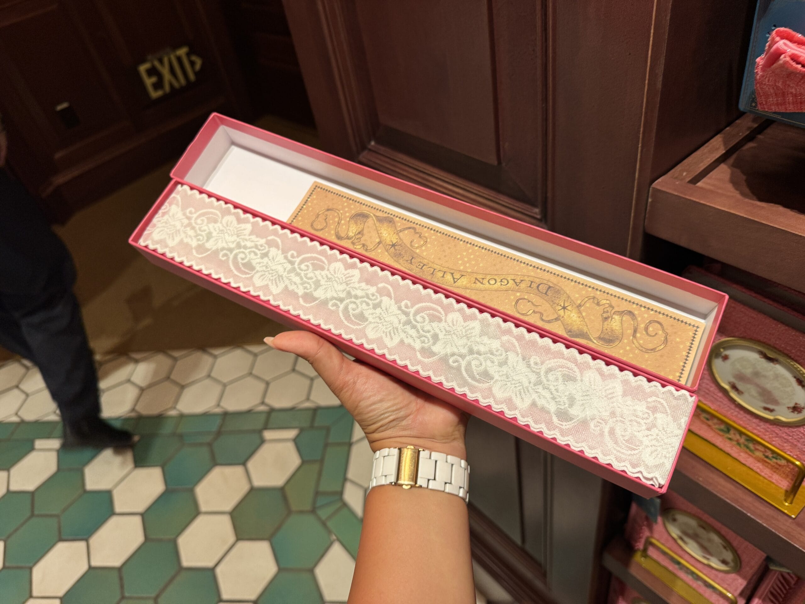 A hand holds an ornate pink box with lace trim containing a Wizarding World merchandise "DIAOON ALLEY" bookmark.
