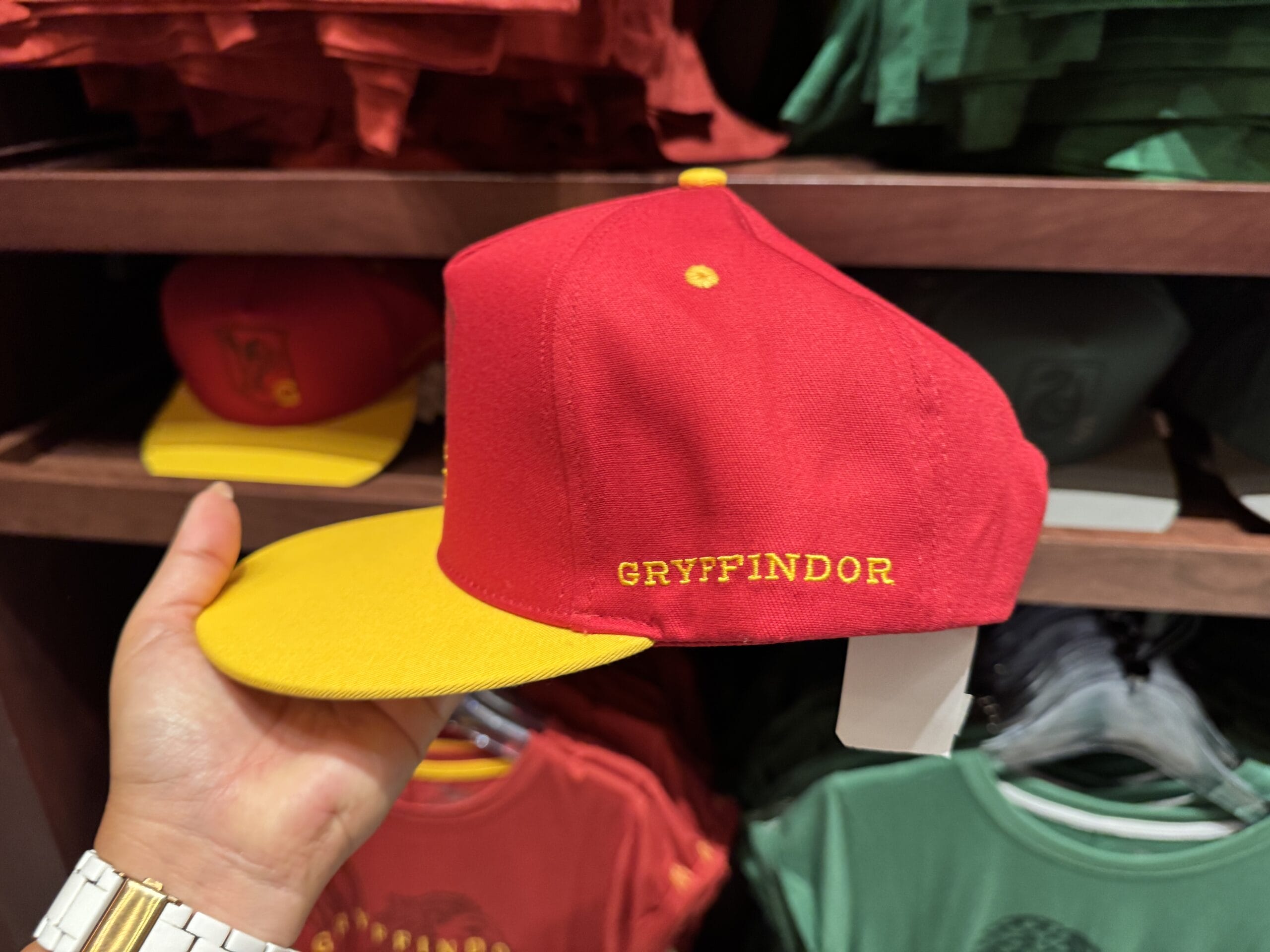 A hand holds a red and yellow cap with "GRYFFINDOR" embroidered—classic Wizarding World Harry Potter merchandise on display.