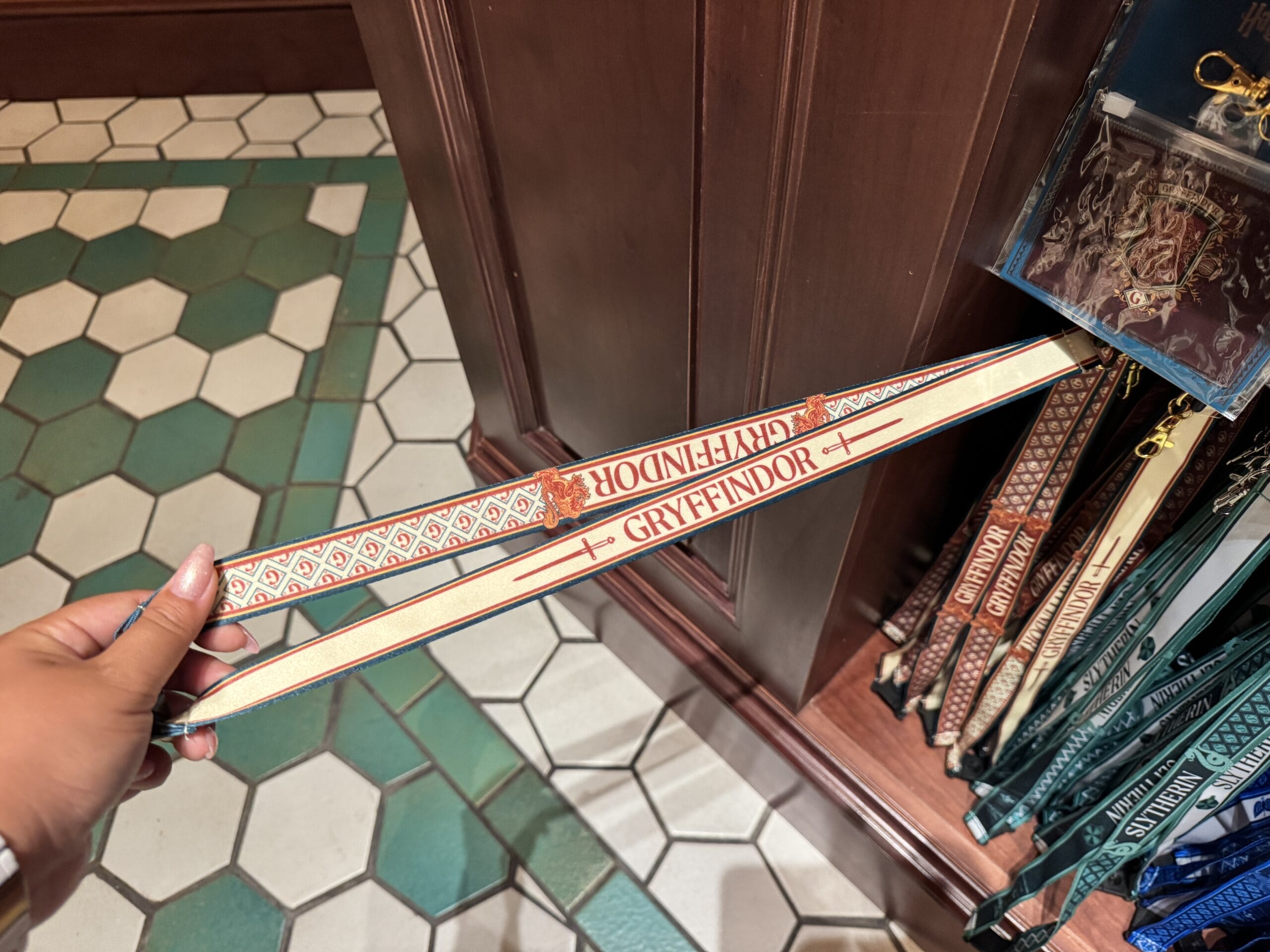 A hand holds a Gryffindor lanyard among Harry Potter merchandise, with other house lanyards hanging in the store.