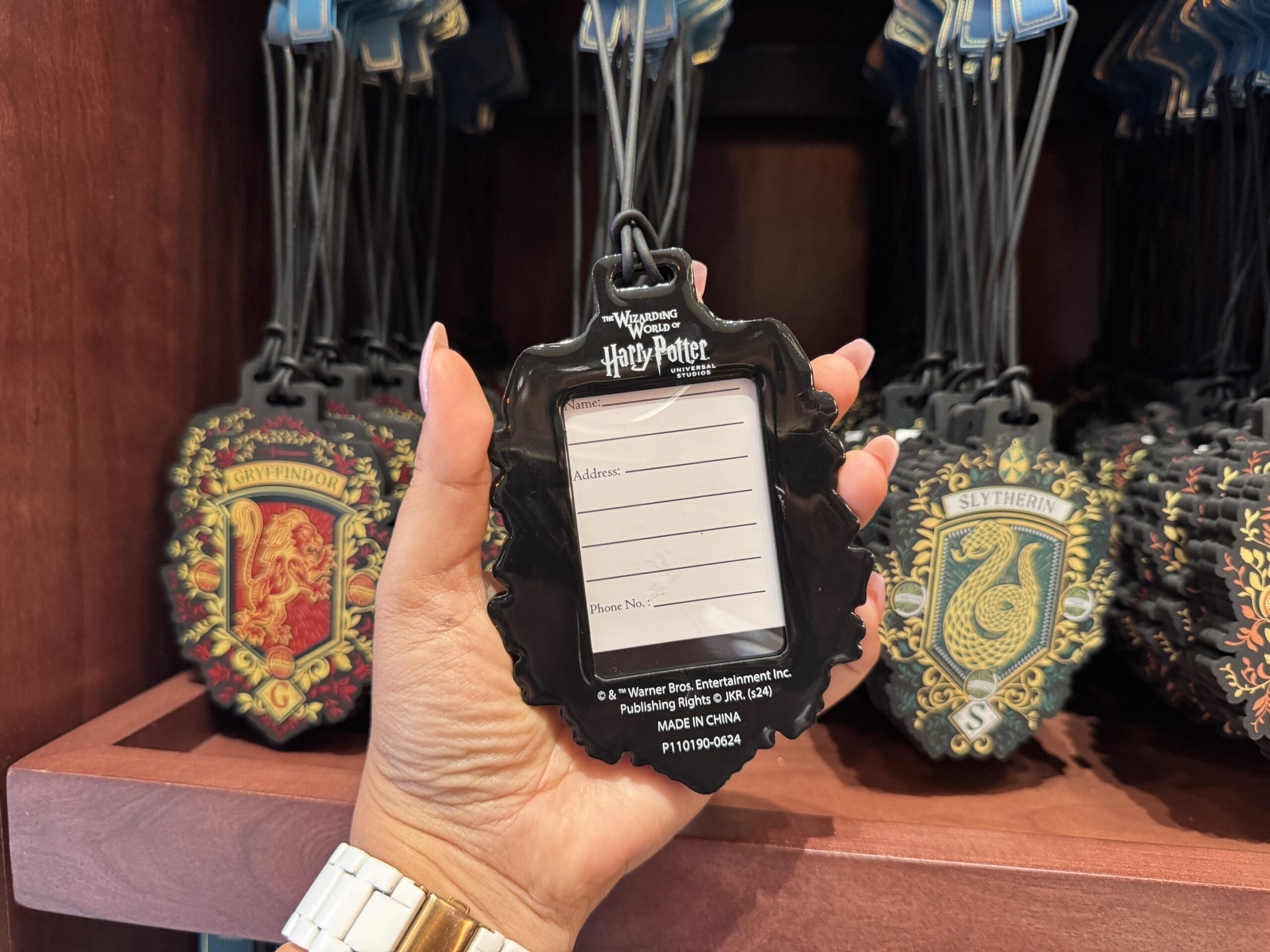 A hand holds a blank Harry Potter-themed luggage tag, part of Wizarding World of Harry Potter merchandise, with house tags behind.