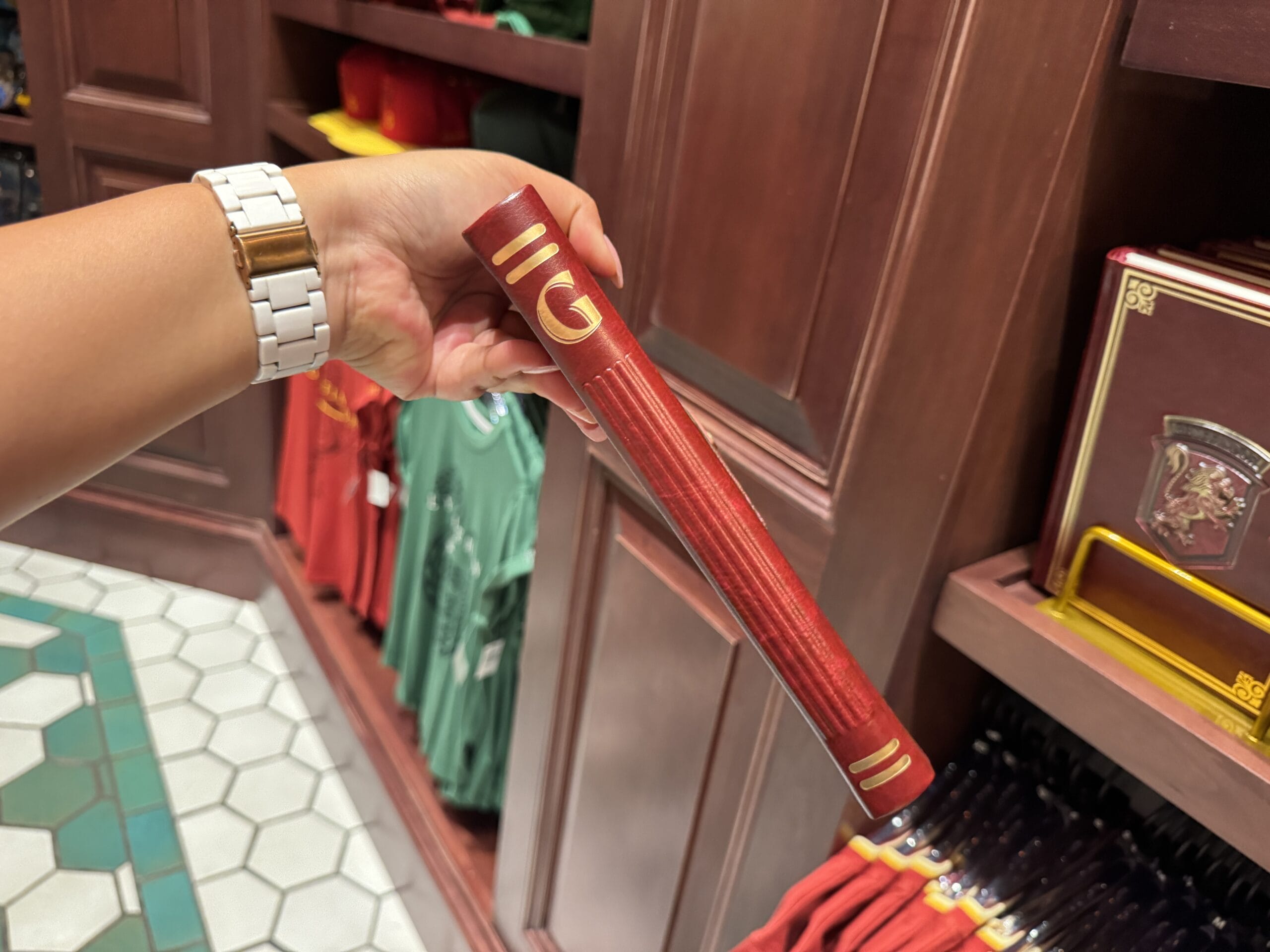 A hand holds a red, wand-shaped object like a book spine with Wizarding World of Harry Potter merchandise on wooden shelves.