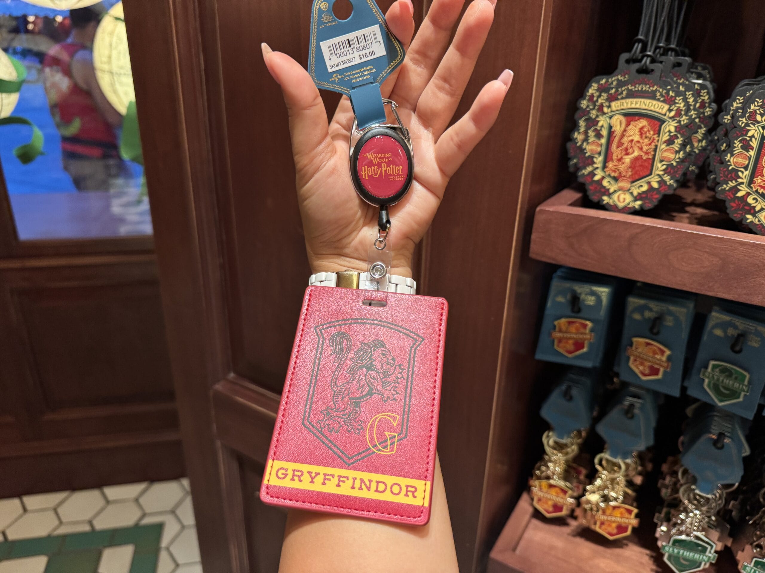 A hand holds a red Gryffindor badge holder from the Wizarding World of Harry Potter merchandise in a store display.