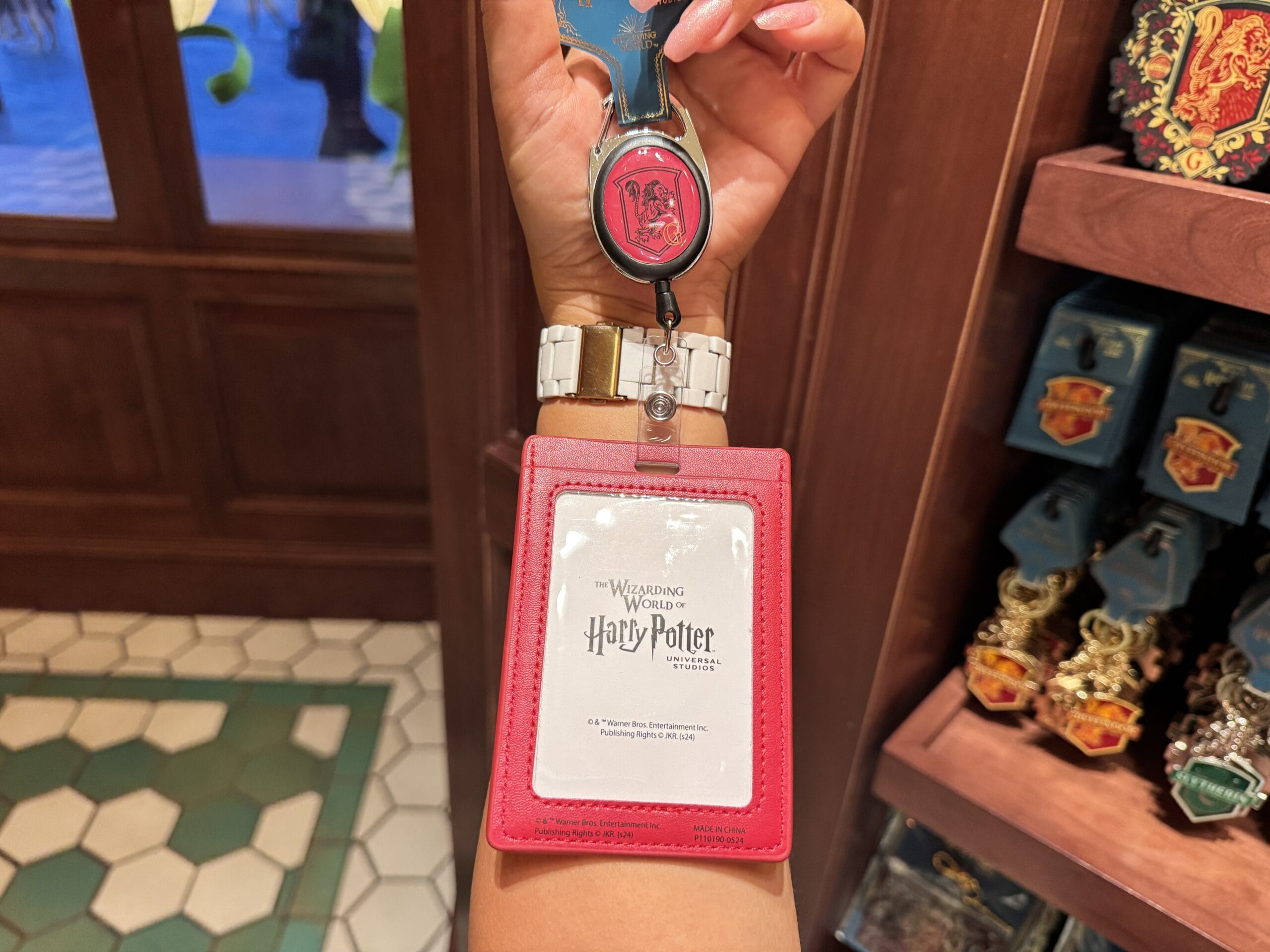 Hand holding a Harry Potter ID badge and Hogwarts keychain among Epic Universe Harry Potter merchandise in-store.