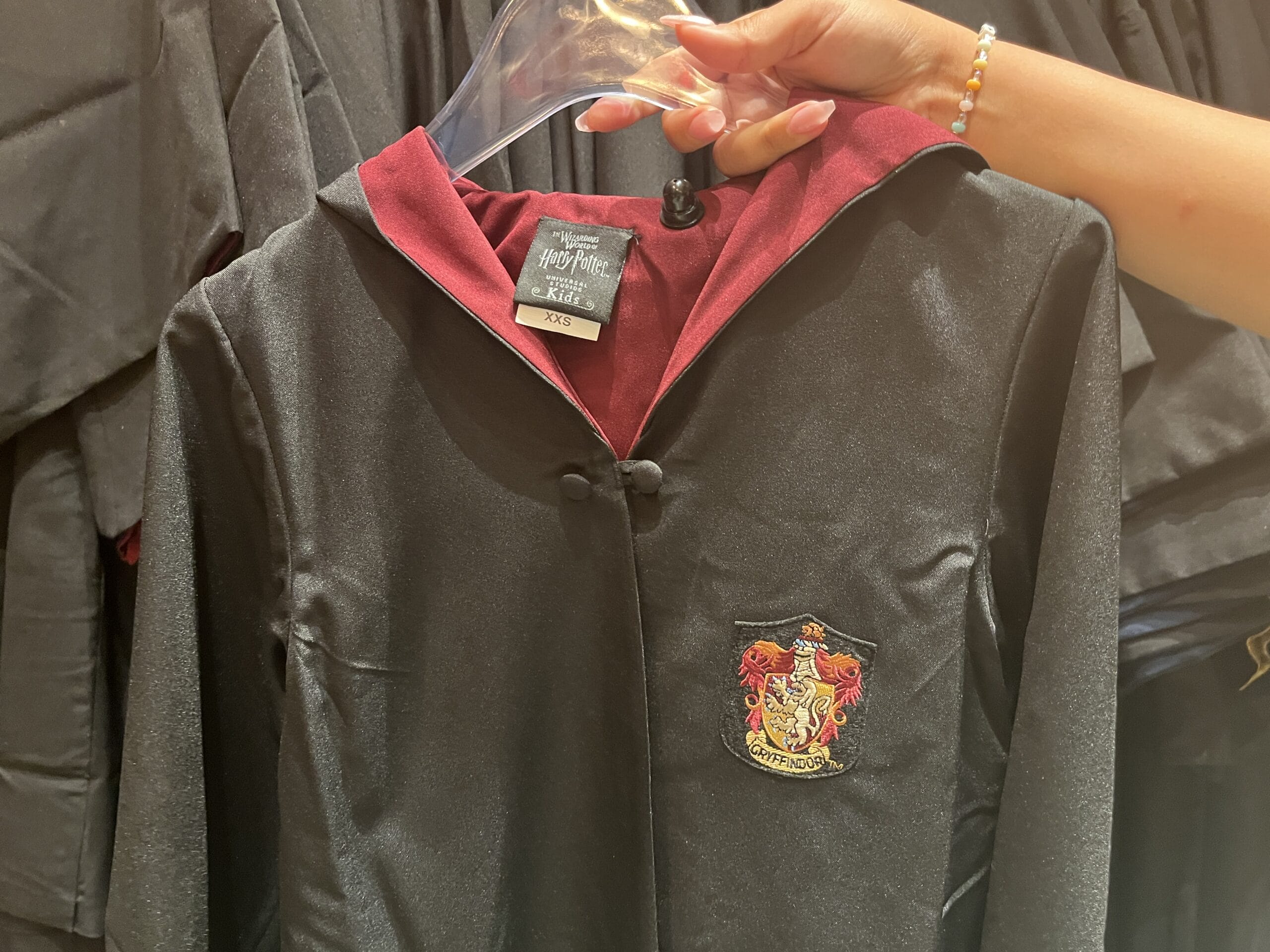 A hand holds a black Gryffindor robe with red lining, Wizarding World Harry Potter merchandise, displayed on a hanger.