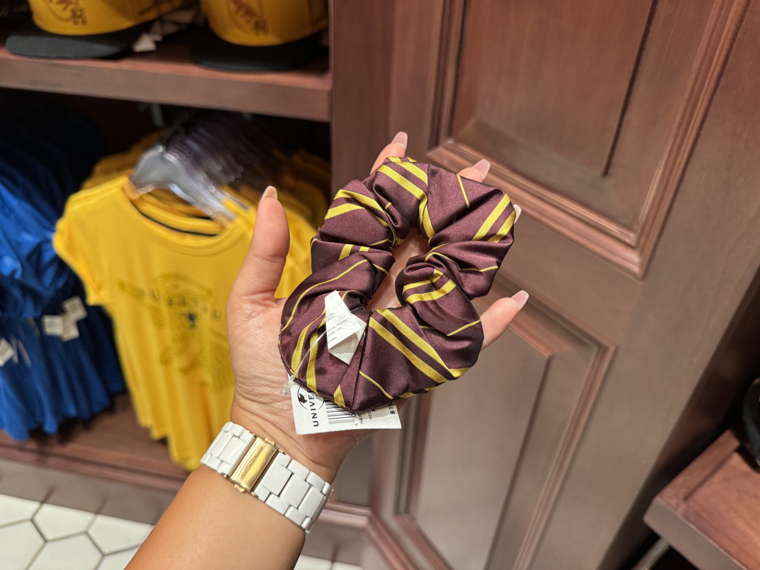 Hand holding a maroon and yellow striped scrunchie, reminiscent of Harry Potter merchandise, with yellow shirts behind.