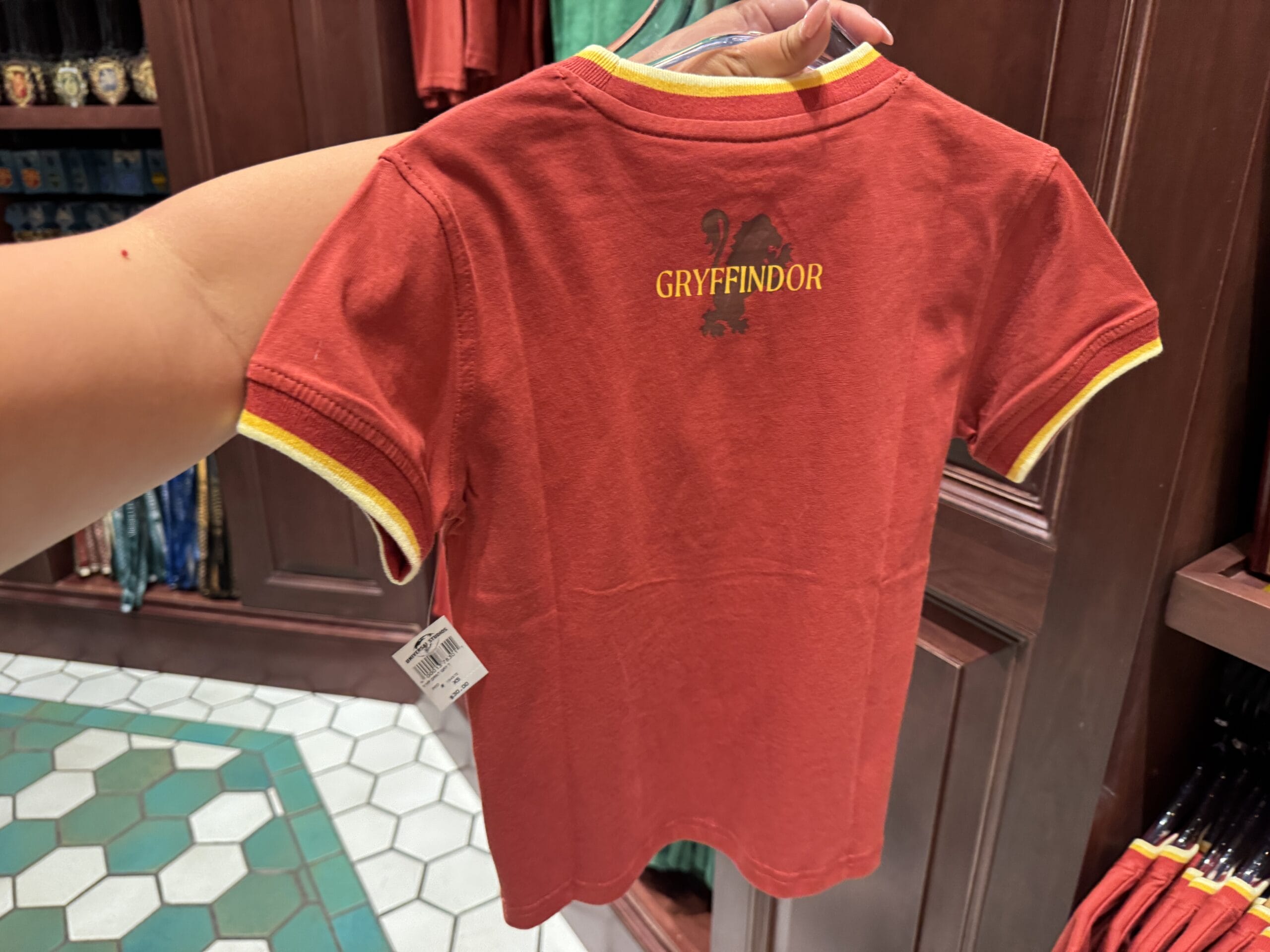 A hand holds a red Gryffindor t-shirt with yellow trim, classic Harry Potter merchandise displayed in a store.