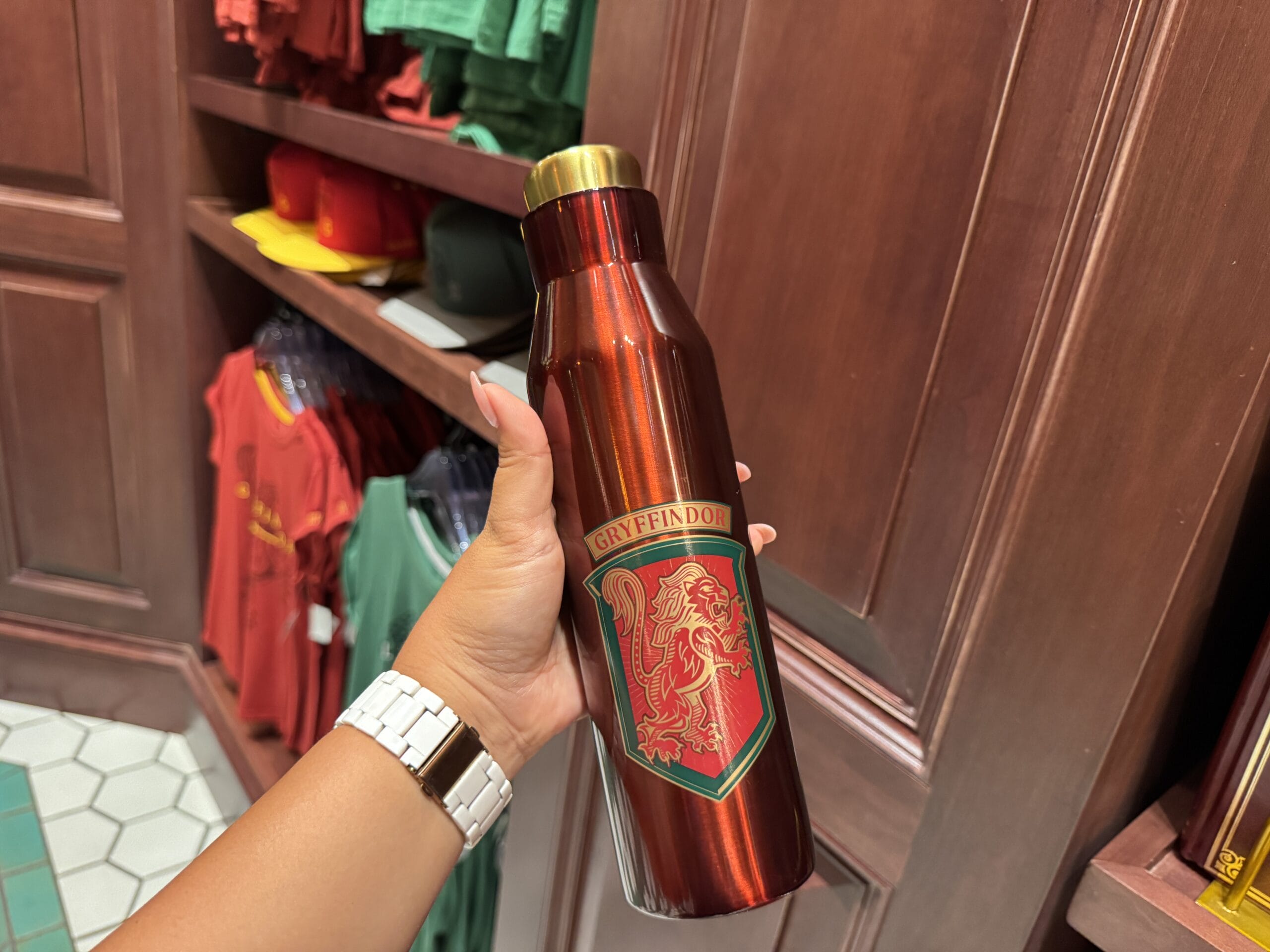 A hand holds a metallic red Gryffindor water bottle amid Wizarding World Harry Potter merchandise in a themed store.