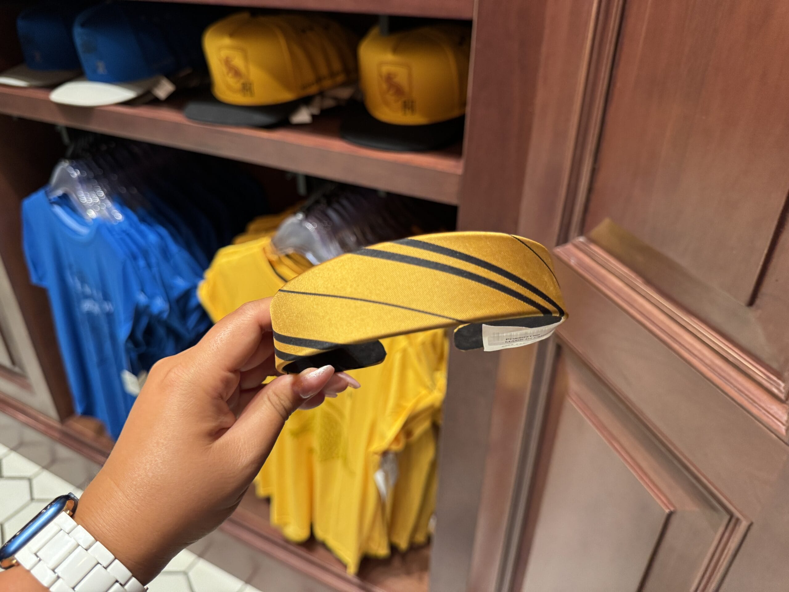A hand holds a yellow headband with black stripes among Wizarding World merchandise in a store filled with yellow hats and shirts.