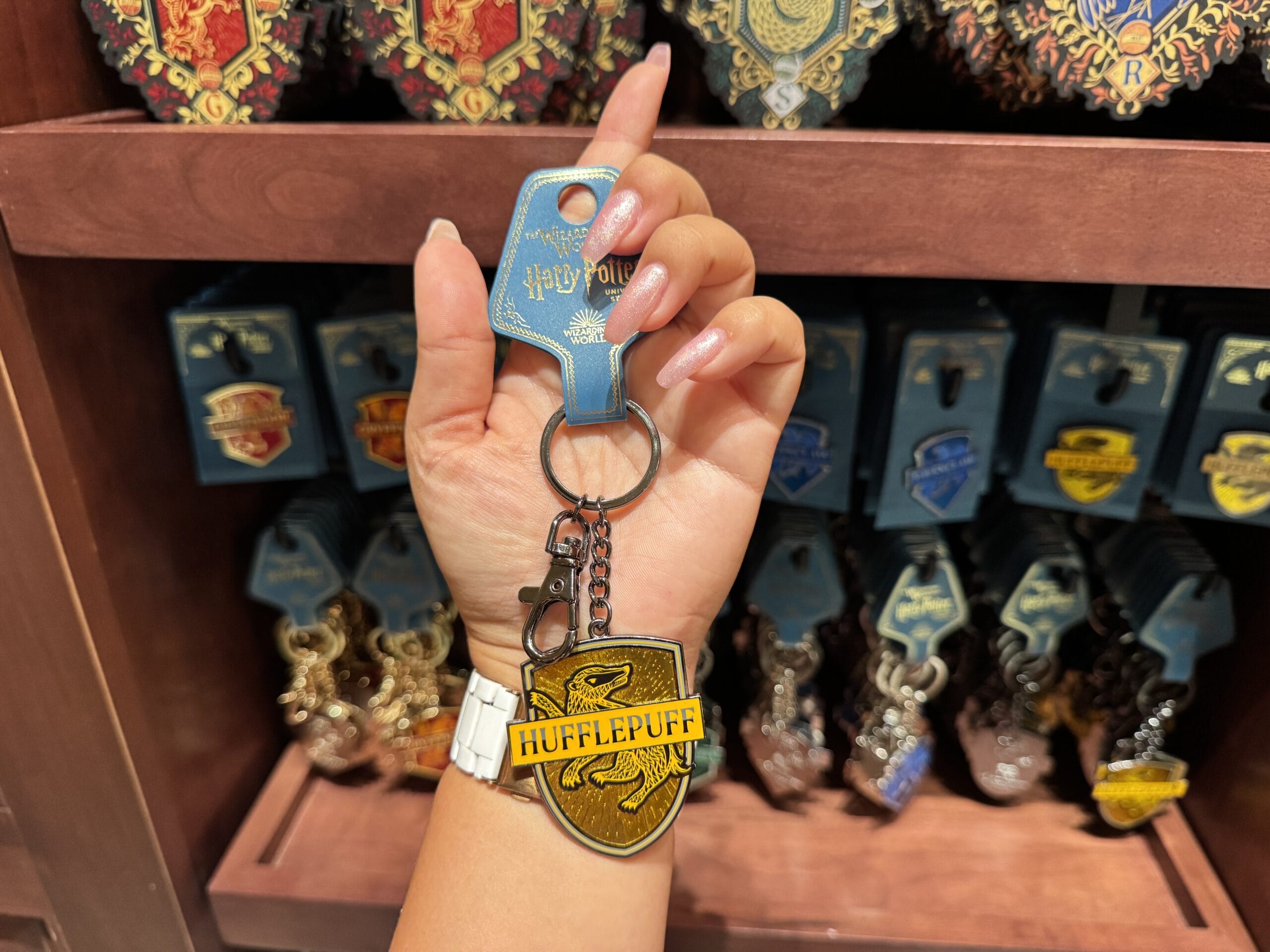 A hand holds a Hufflepuff keychain in a store, surrounded by Harry Potter merchandise in the background.