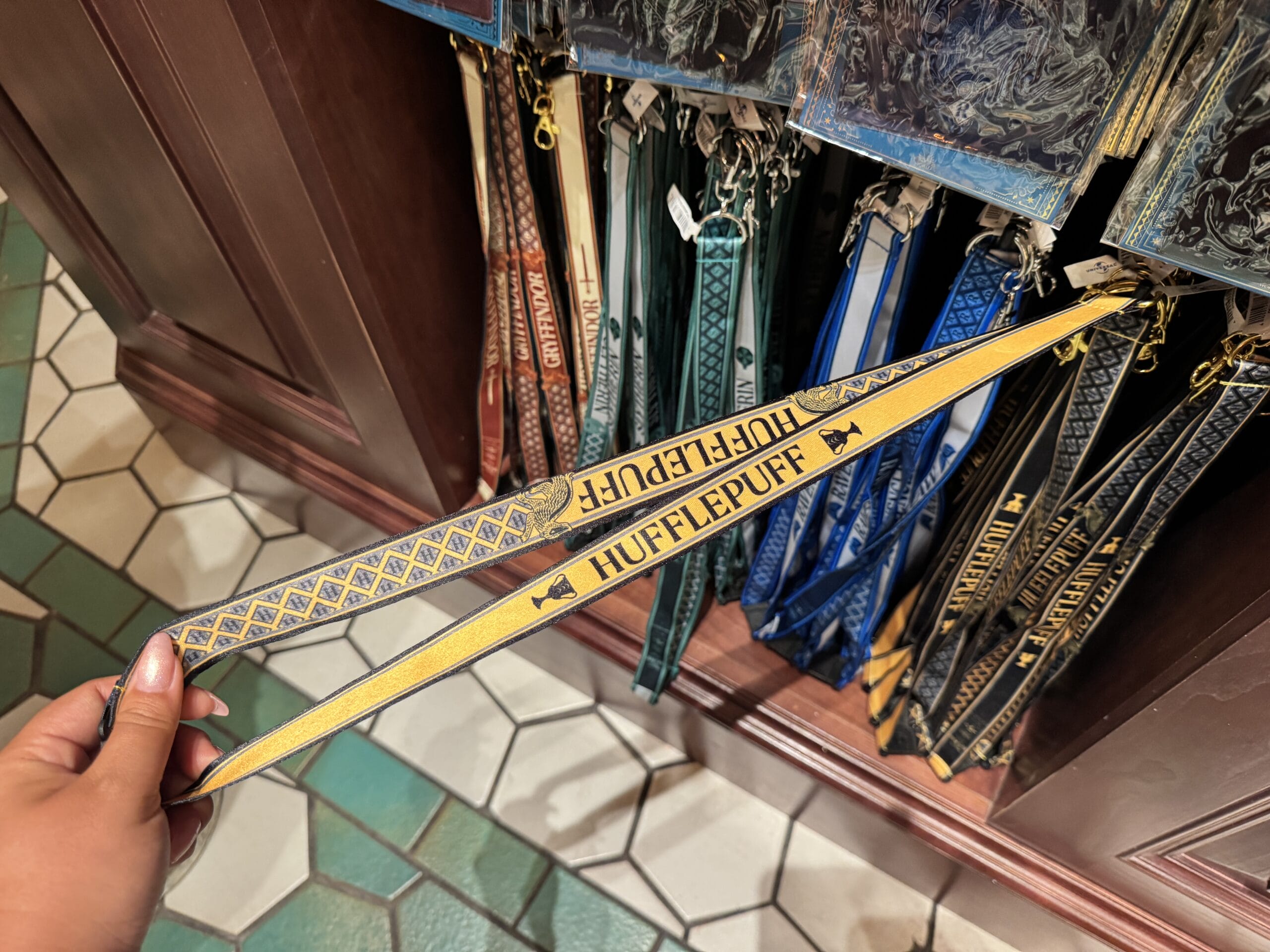 A hand holds a yellow Hufflepuff lanyard with other Harry Potter house lanyards in Wizarding World merchandise display.