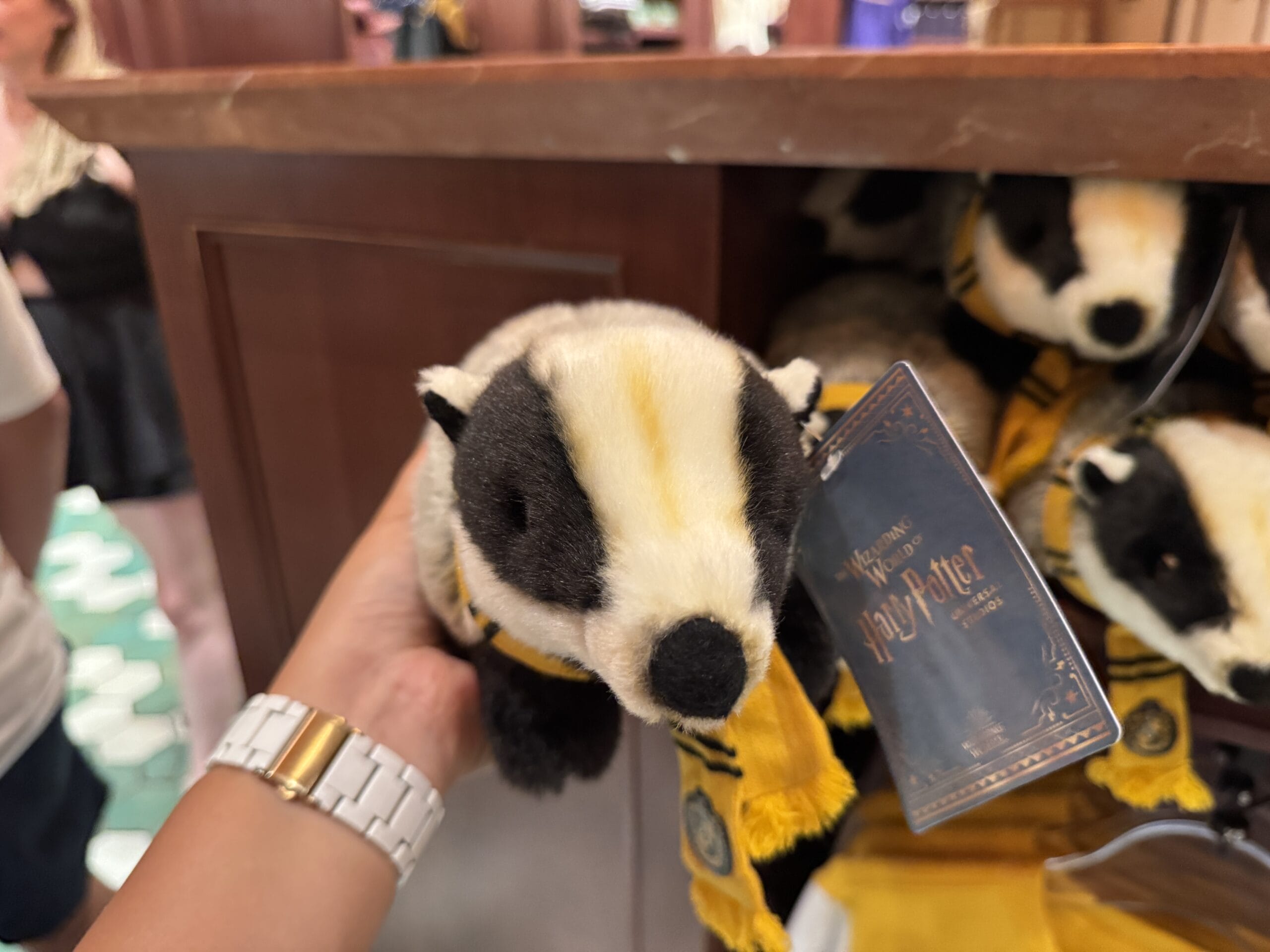 A hand holds a plush badger toy with a yellow scarf at the Wizarding World of Harry Potter merchandise store.