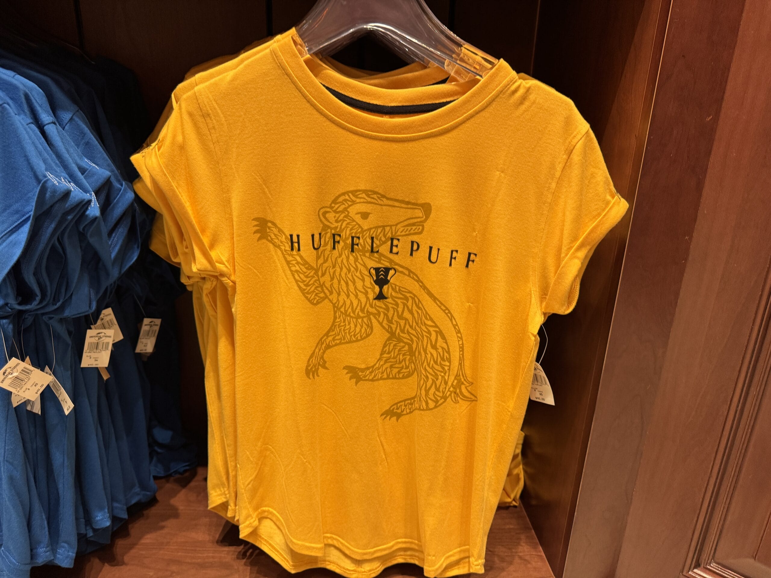 Yellow Hufflepuff t-shirt with a badger graphic, official Wizarding World of Harry Potter merchandise, on rack by blue shirts.