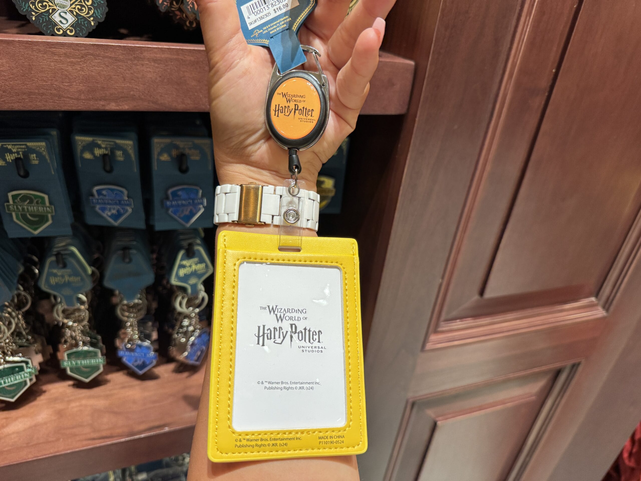 A hand holds Wizarding World merchandise—a yellow Harry Potter ID holder and retractable badge clip—in a gift shop.