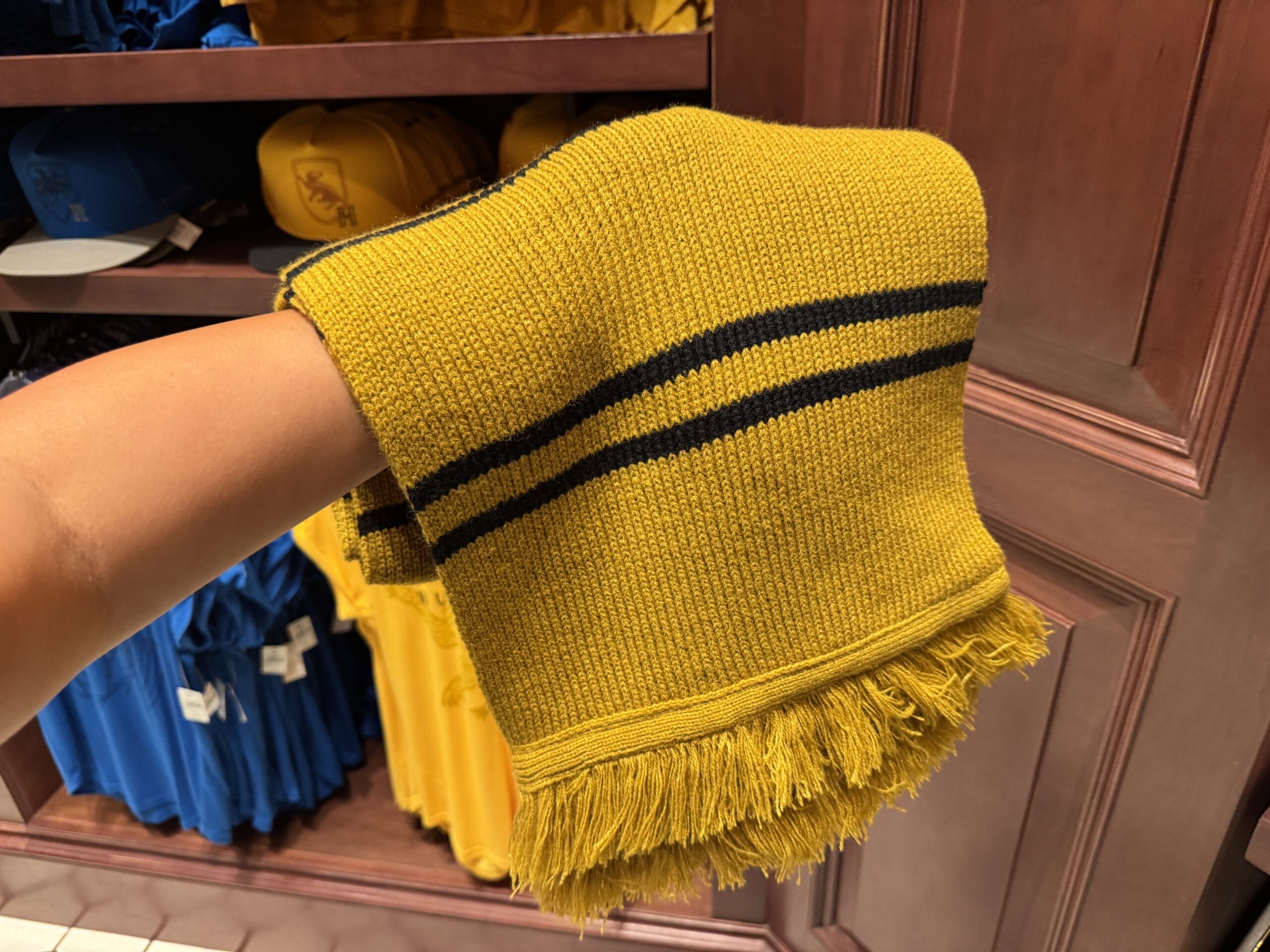 A hand holds Wizarding World of Harry Potter merchandise—a yellow, black-striped scarf—before shelves of folded clothes.