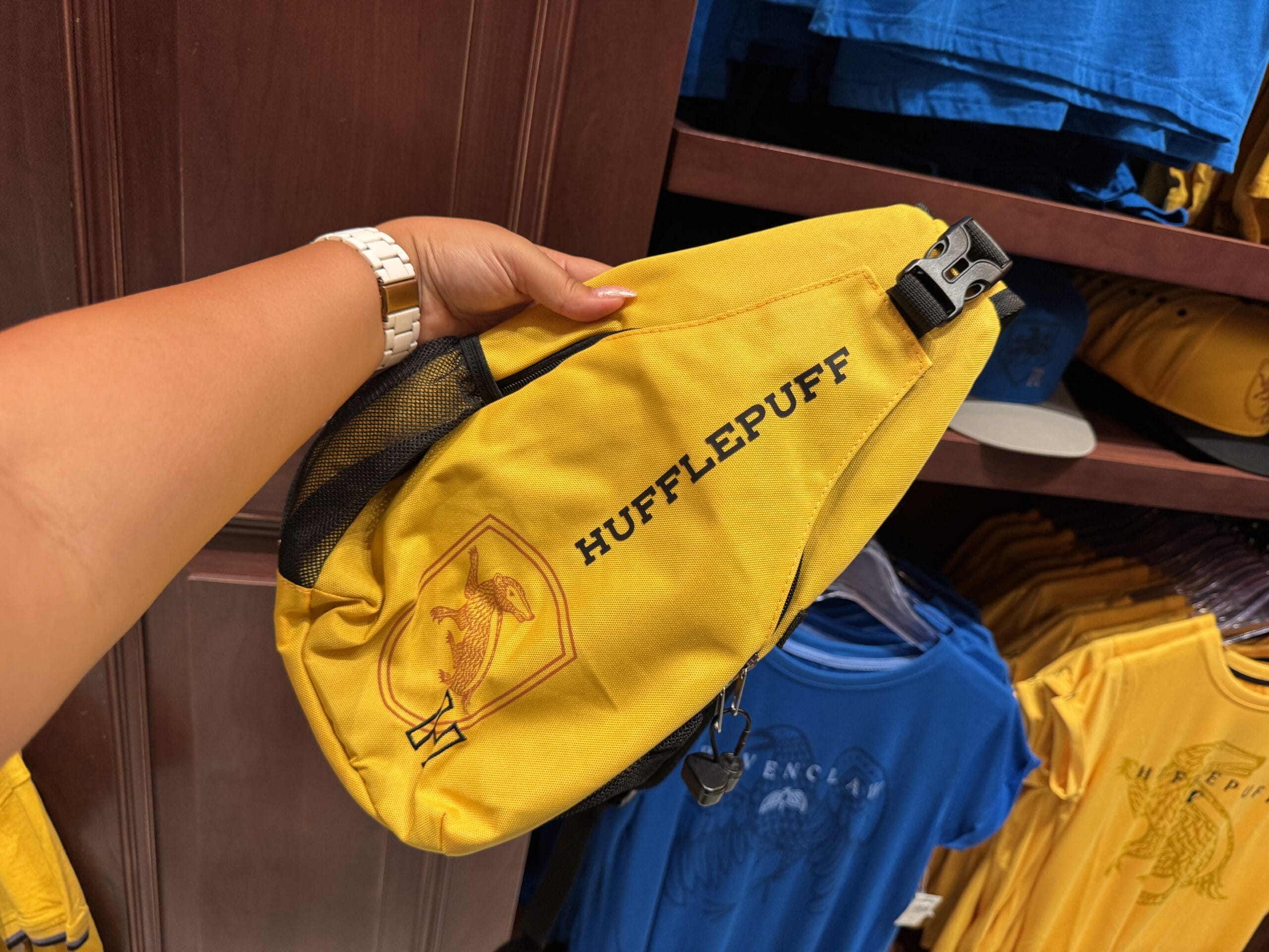 A hand holds a yellow Hufflepuff sling bag with crest, near shirts on display—perfect Epic Universe merchandise.