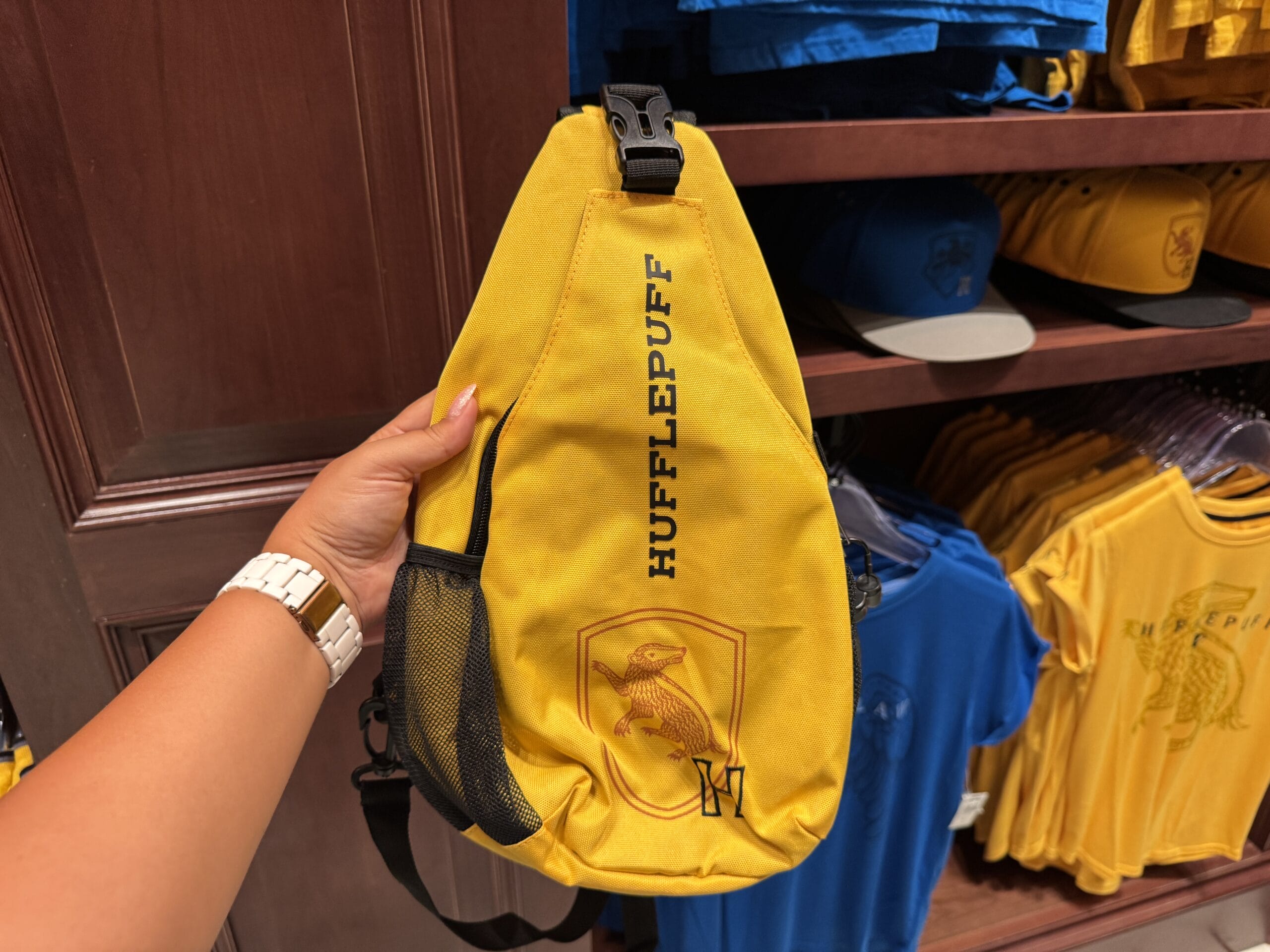 A hand holds a yellow Hufflepuff sling bag with a badger emblem, seen among Wizarding World merchandise in a store.