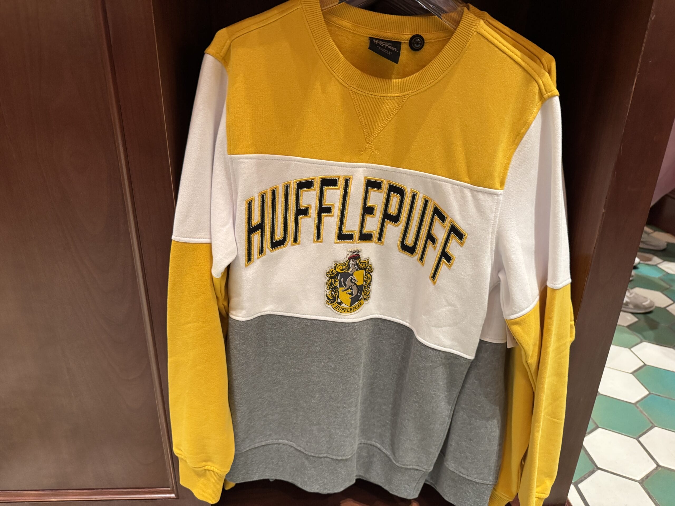 A yellow, white, and gray Hufflepuff sweatshirt—official Wizarding World of Harry Potter merchandise—hangs on a wooden rack.