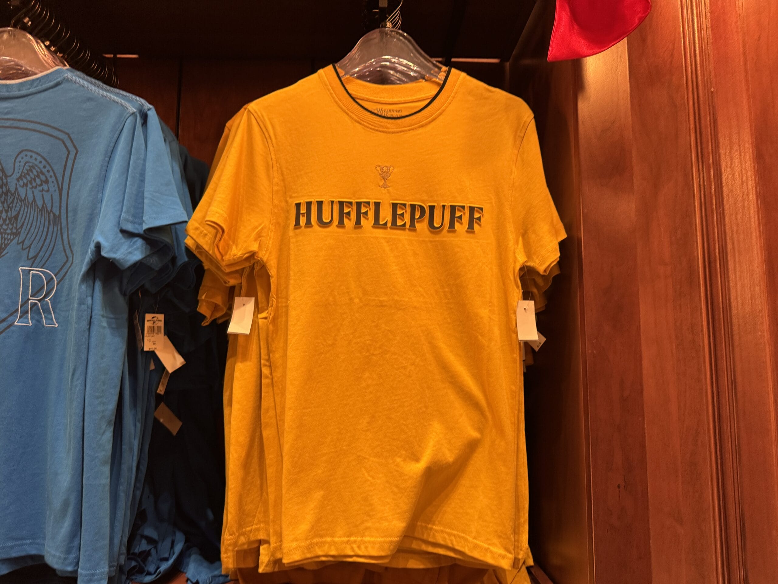 A yellow Hufflepuff T-shirt hangs in-store beside a blue shirt—perfect Wizarding World of Harry Potter merchandise.