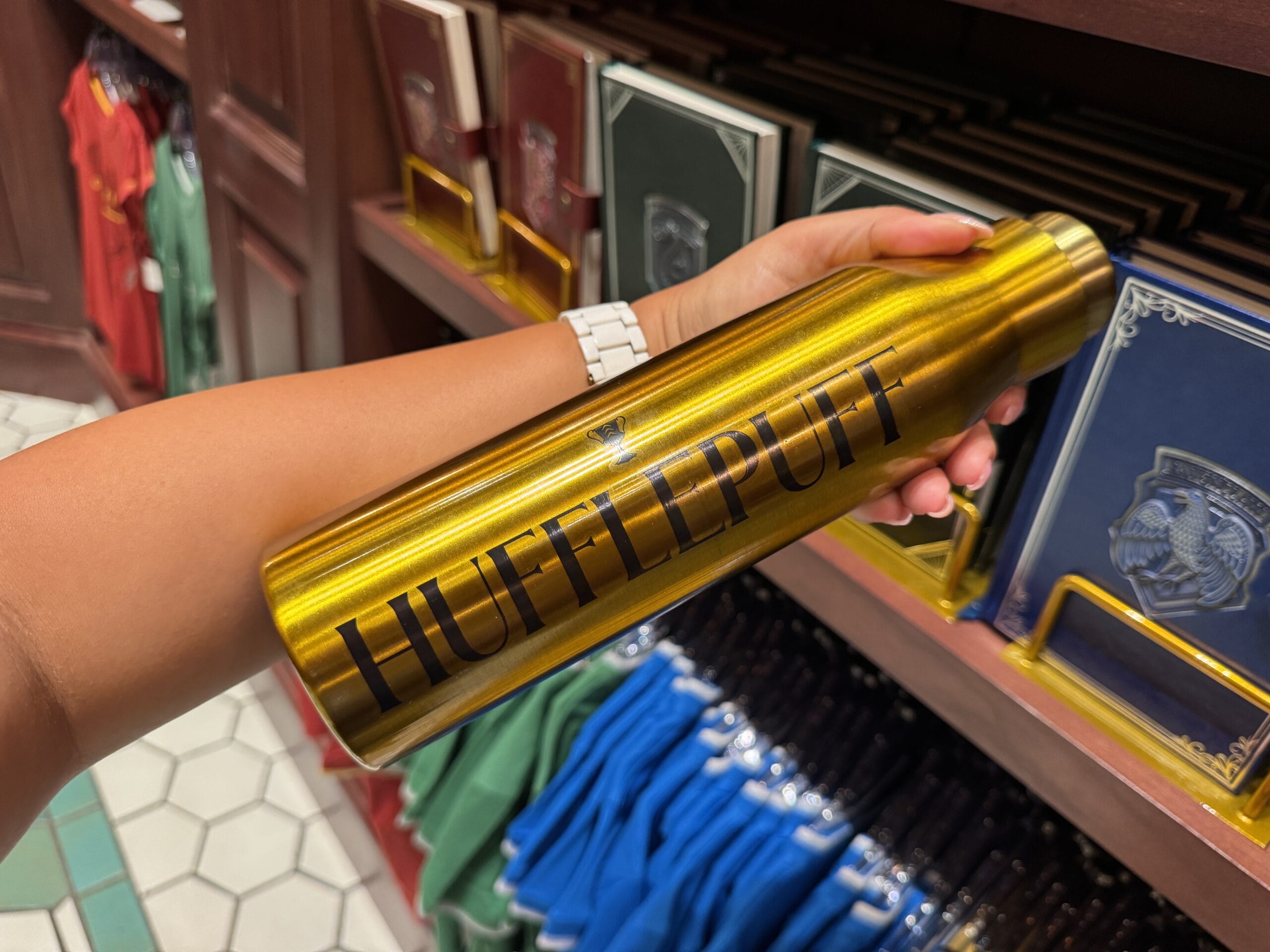 A person holds a shiny gold "Hufflepuff" water bottle among Wizarding World and Harry Potter merchandise in a store.