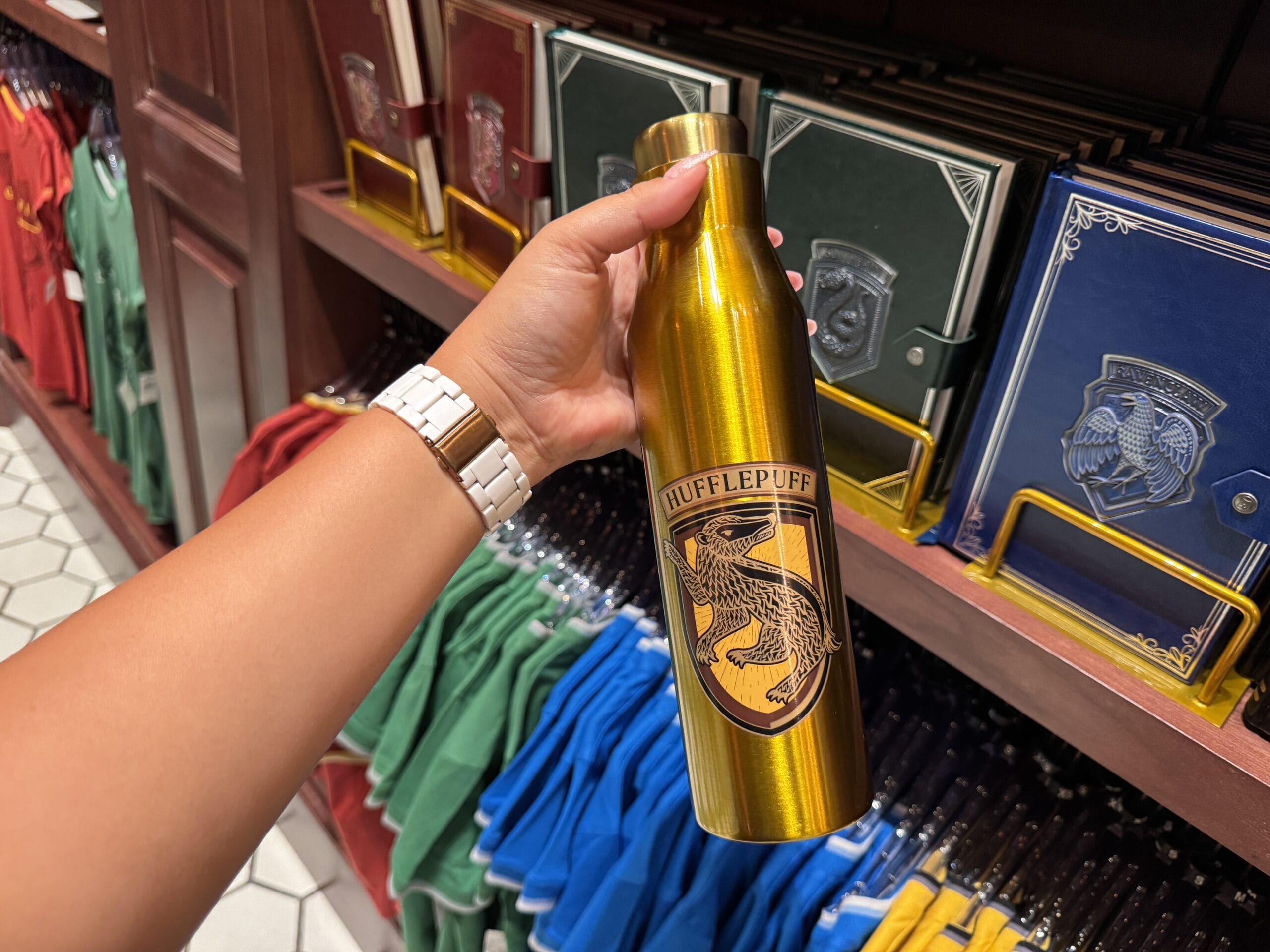A hand holds a gold Hufflepuff water bottle in a shop filled with Wizarding World merchandise and Hogwarts house notebooks.