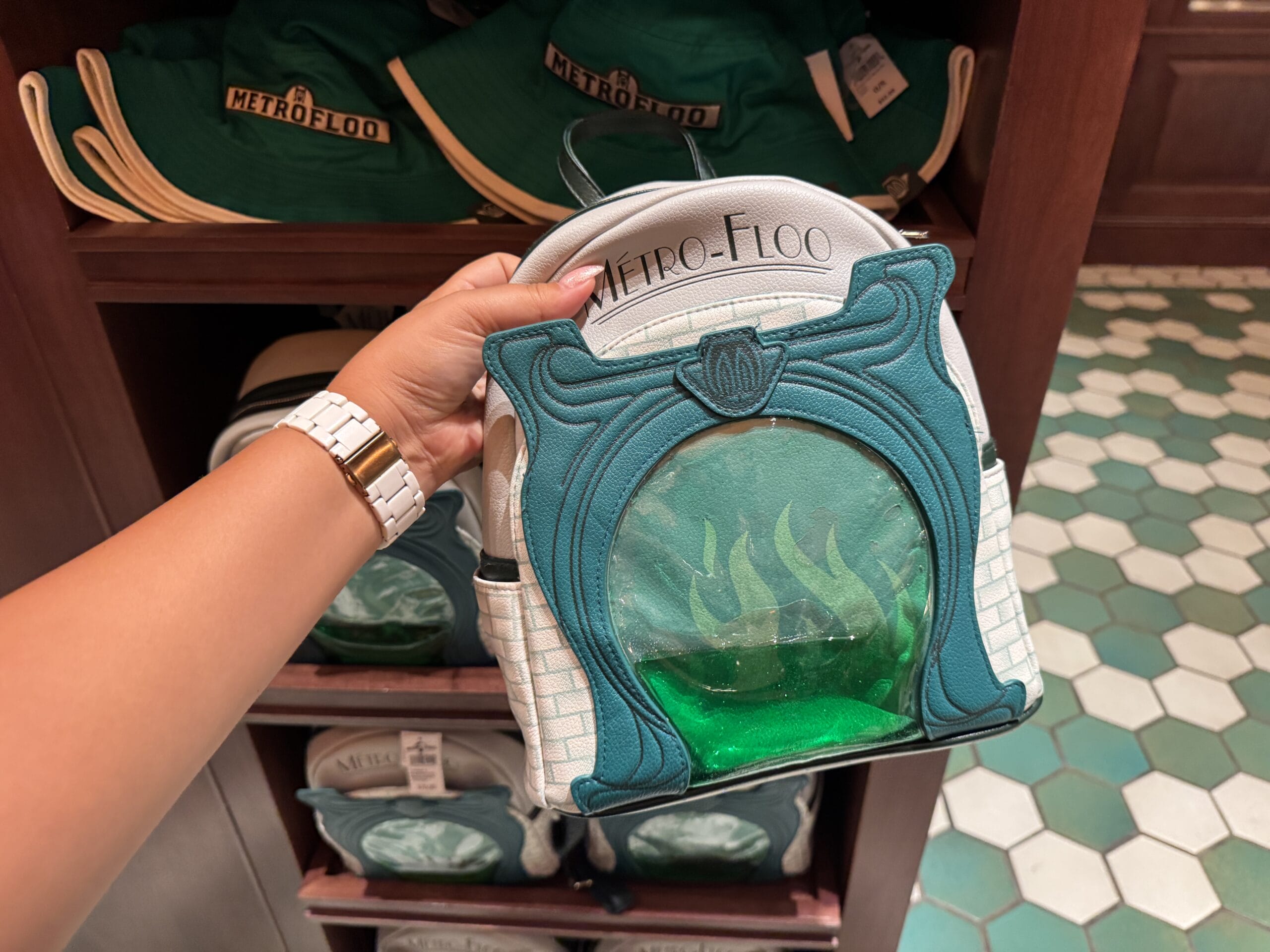 A hand holds a Metro-Floo themed bag with green flames, surrounded by Epic Universe hats and Harry Potter merchandise.