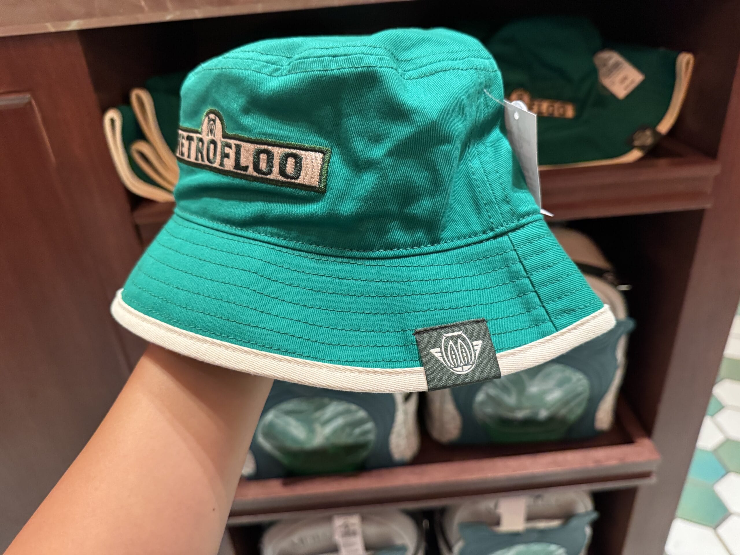 A hand holds a green bucket hat with a "RETROFLOO" patch, inspired by Tour En Floo, on a store display.