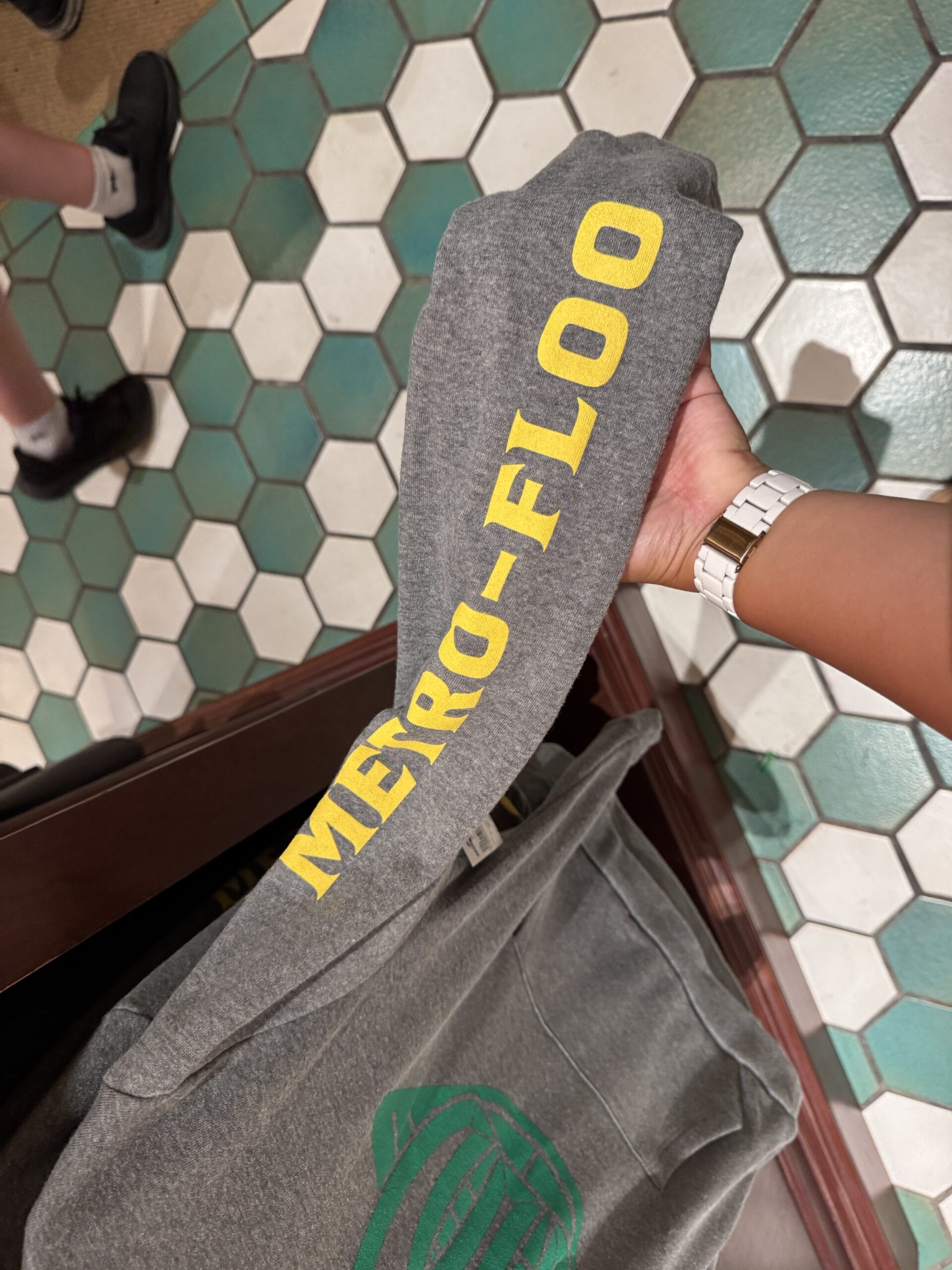 A hand displays METRO-FLOO merchandise over tiled green and white floors at Epic Universe.
