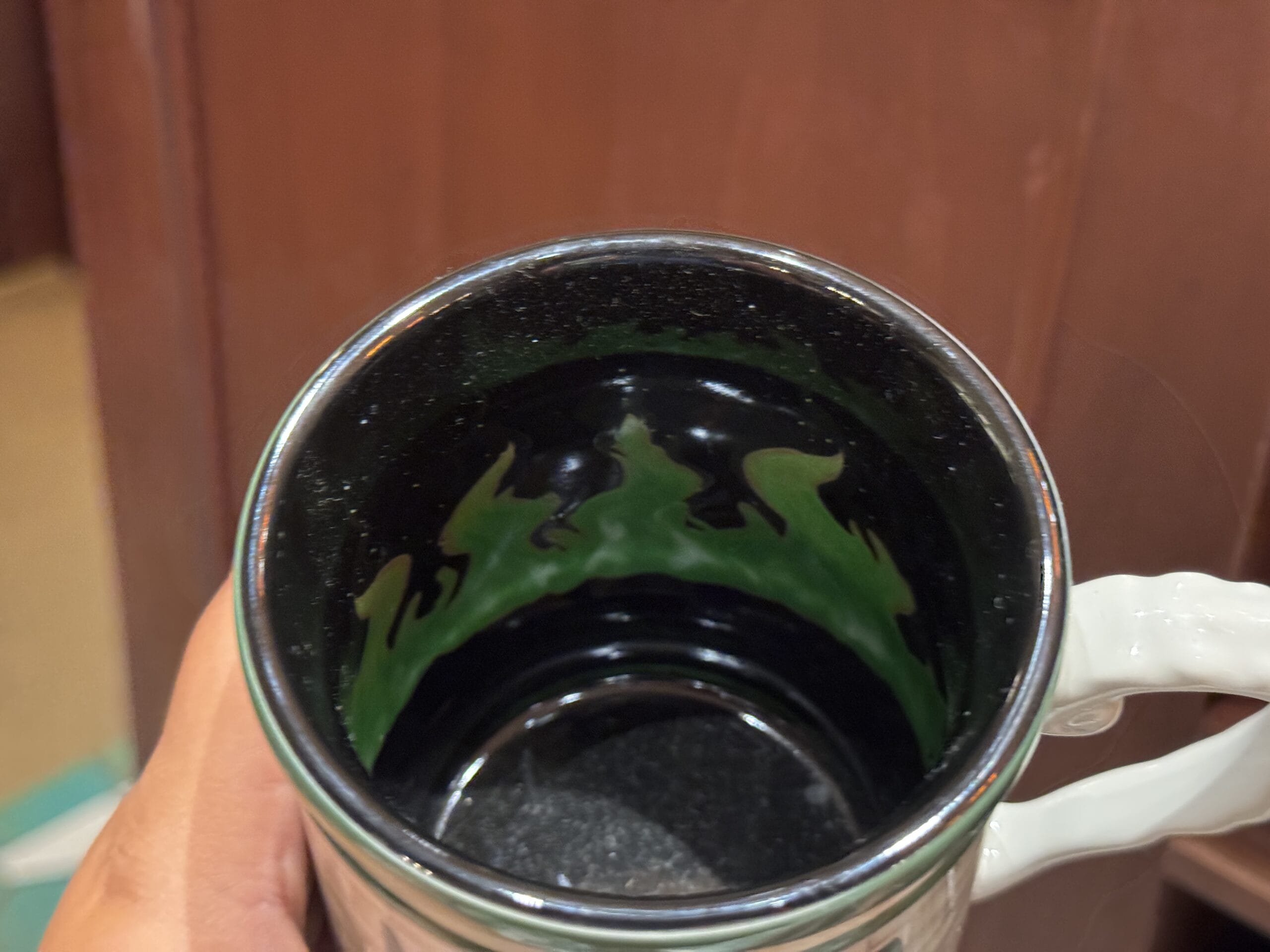 A hand holds a ceramic mug with a black and green pattern, perfect Wizarding World merchandise, in front of a wooden background.