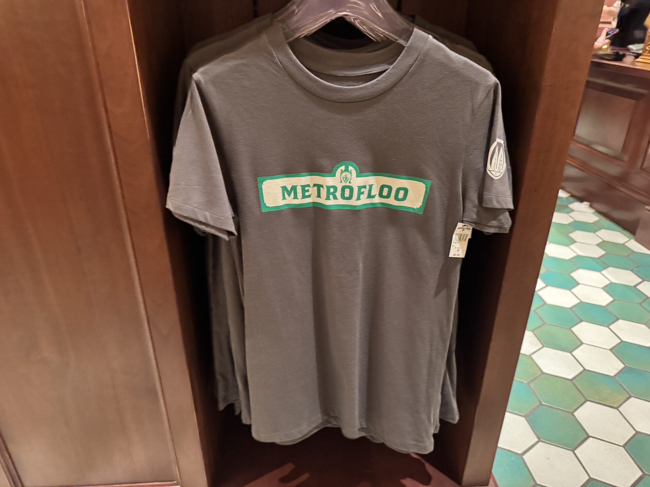 Gray t-shirt with "METROFLOO" in green, inspired by Tour En Floo at Epic Universe, displayed on a wooden rack in-store.