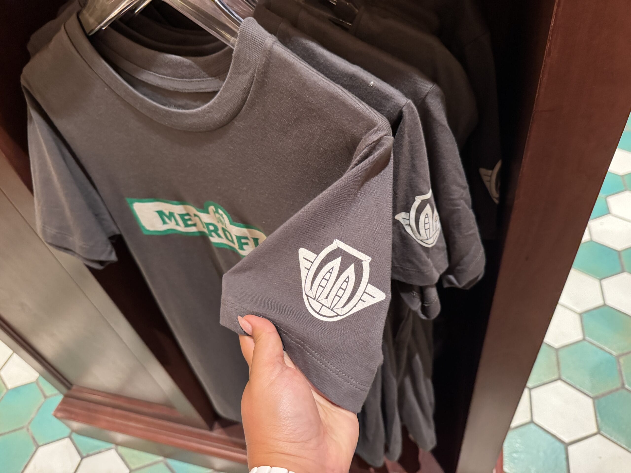 A hand holds MERCEDES merchandise—a dark gray T-shirt with a white logo—displayed on a clothing rack.