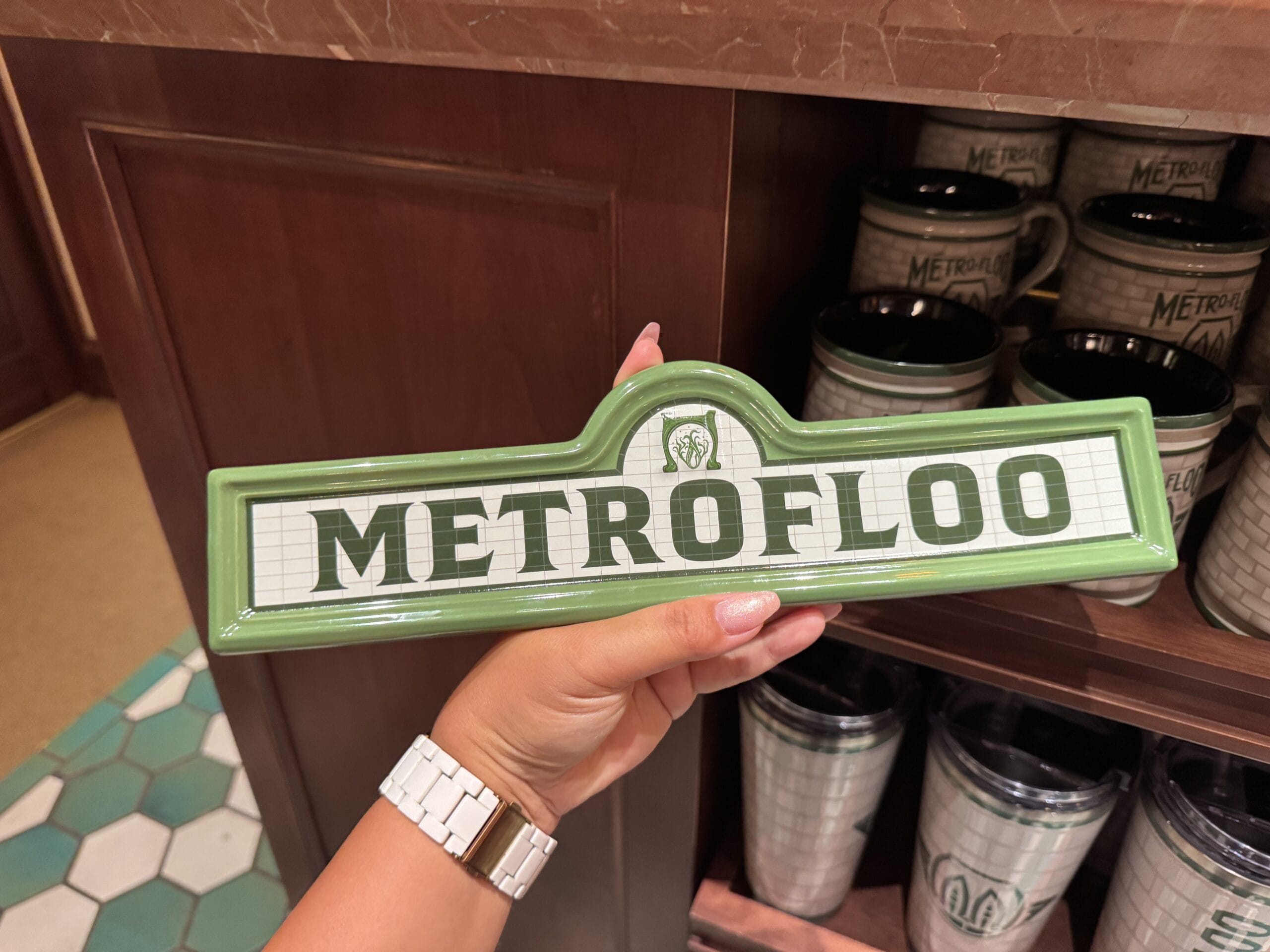 A hand holds a green sign reading “METROFLOO” in front of shelves with Wizarding World merchandise and mugs.