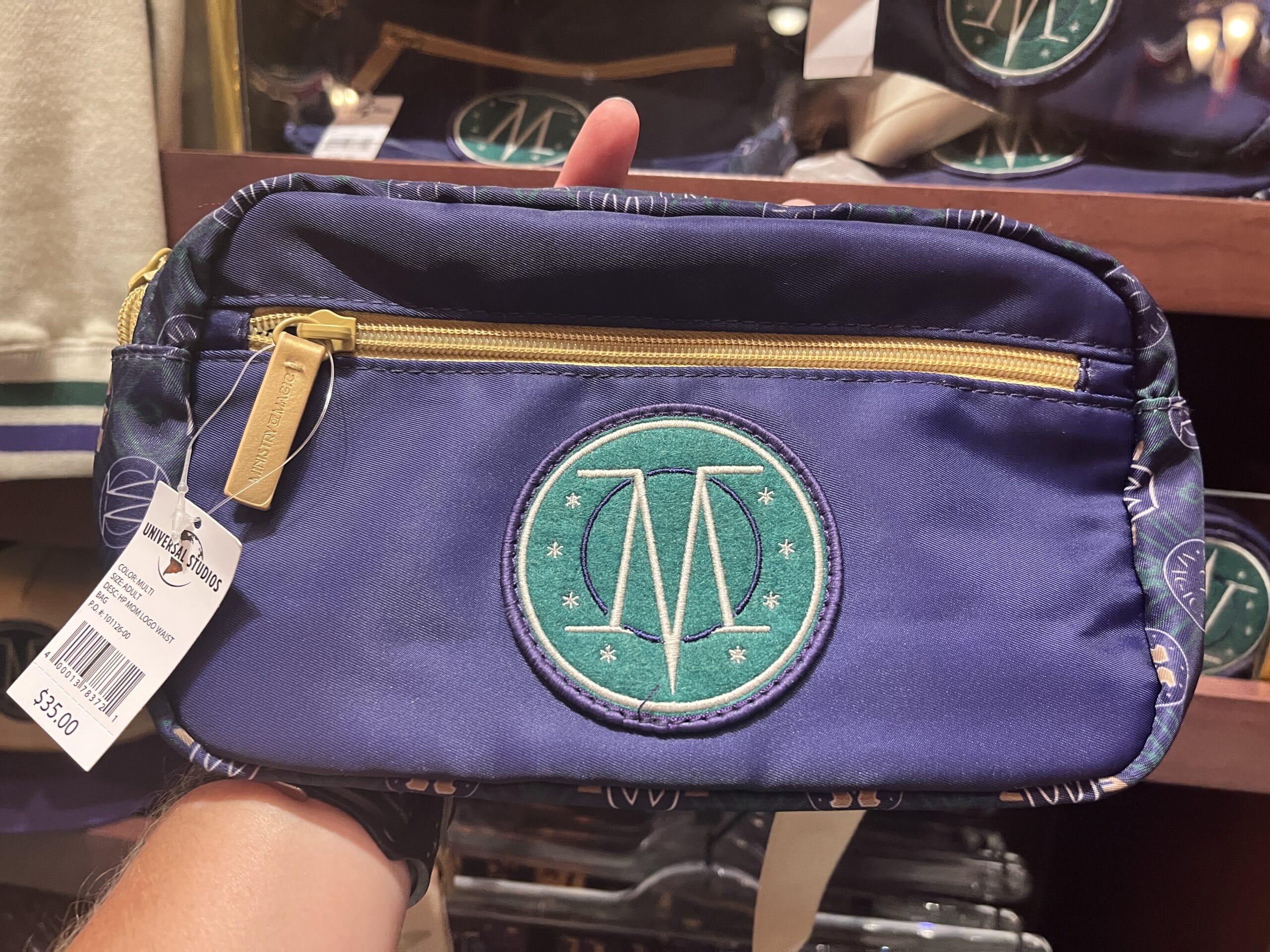 A hand holds Wizarding World merchandise: a blue pouch with gold zipper, green "TM" emblem, and $35 price tag.