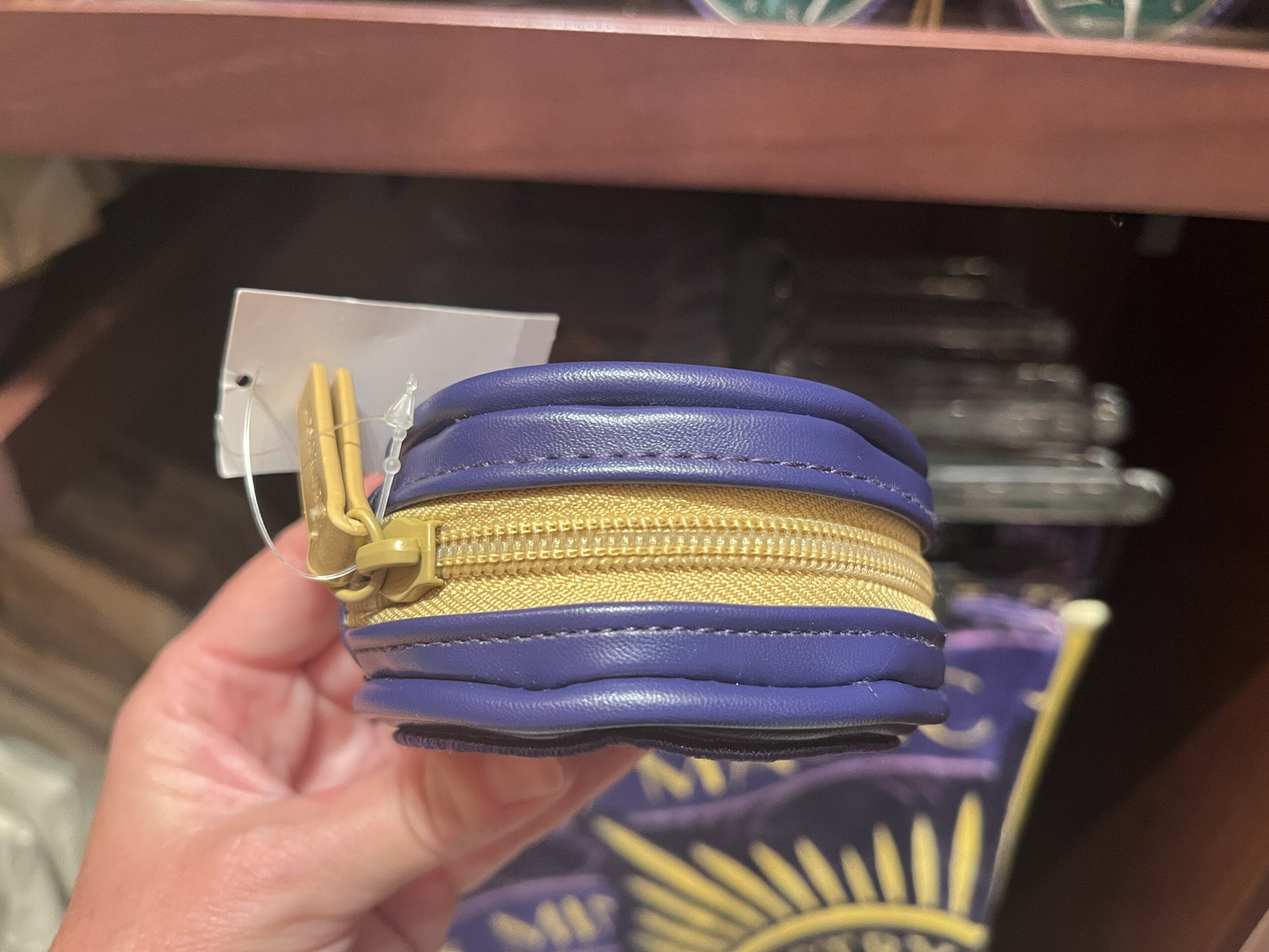 A hand holds Epic Universe merchandise: a small blue pouch with a yellow zipper and a white tag attached.