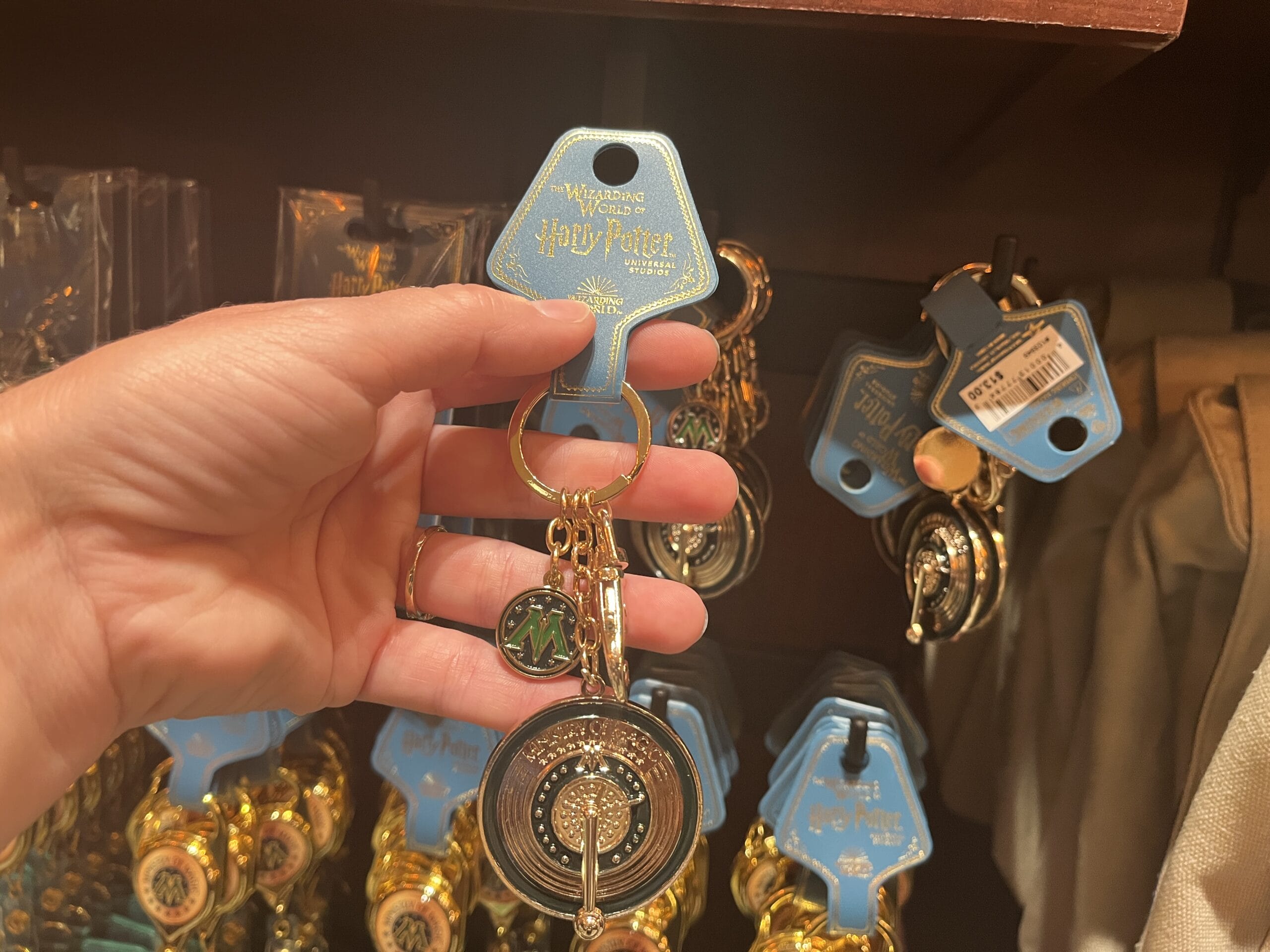 A hand holds Wizarding World of Harry Potter merchandise: a time turner keychain with green "M" charm on display.