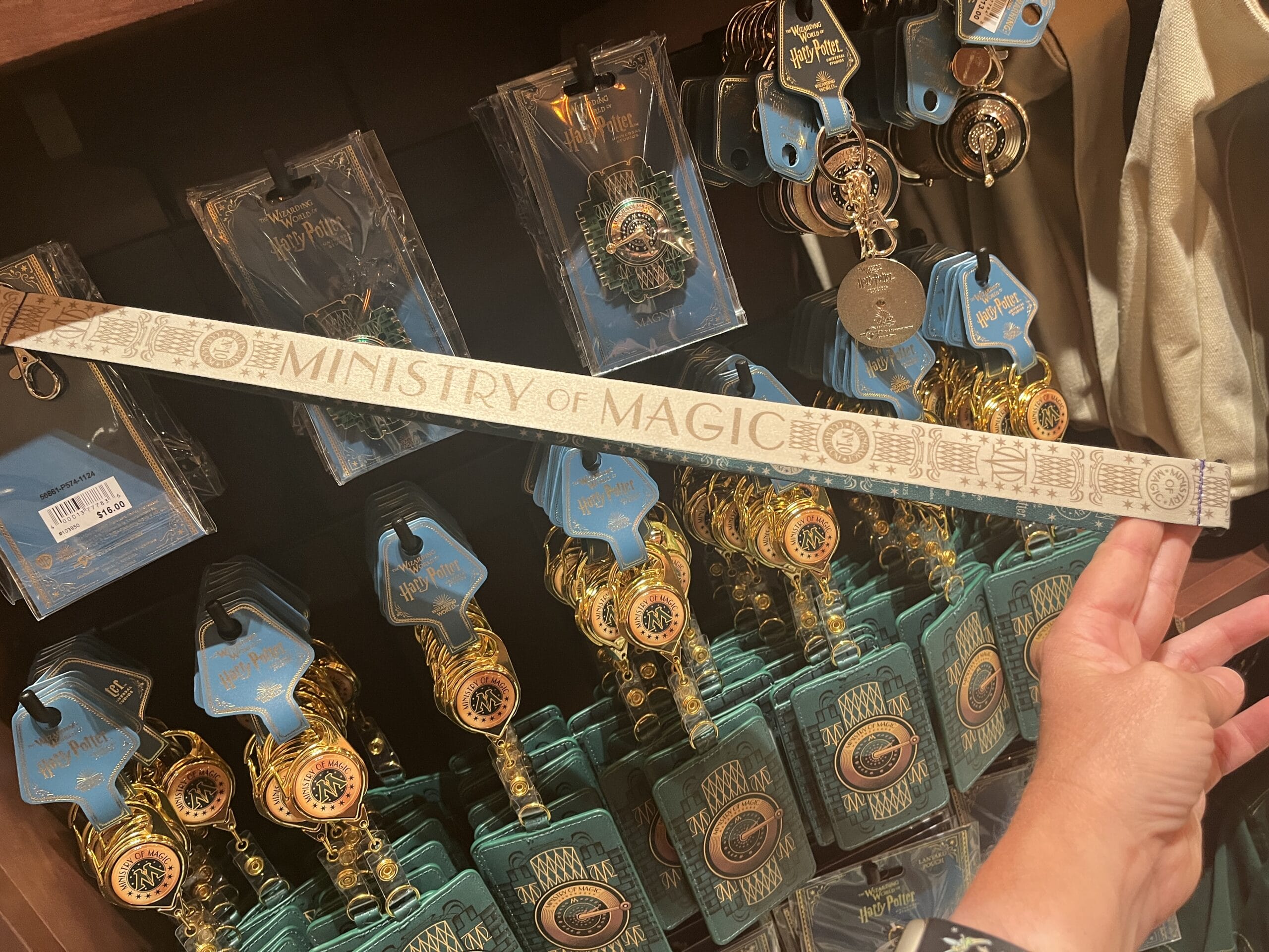 A hand holds a "Ministry of Magic" lanyard before Wizarding World of Harry Potter merchandise on display.