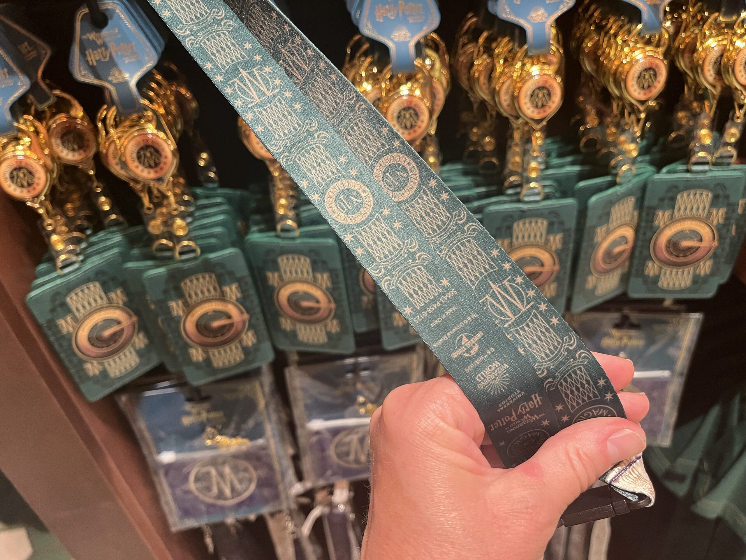 A hand holds a green lanyard with gold Hogwarts icons amid Wizarding World of Harry Potter merchandise on display.