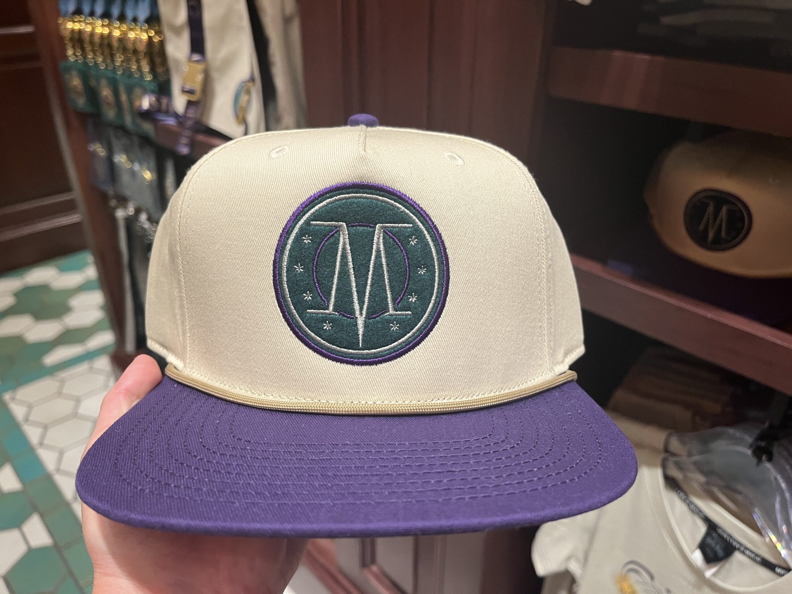 A hand holds a beige and purple hat with a green "M" logo, perfect Wizarding World merchandise for your Harry Potter tour.