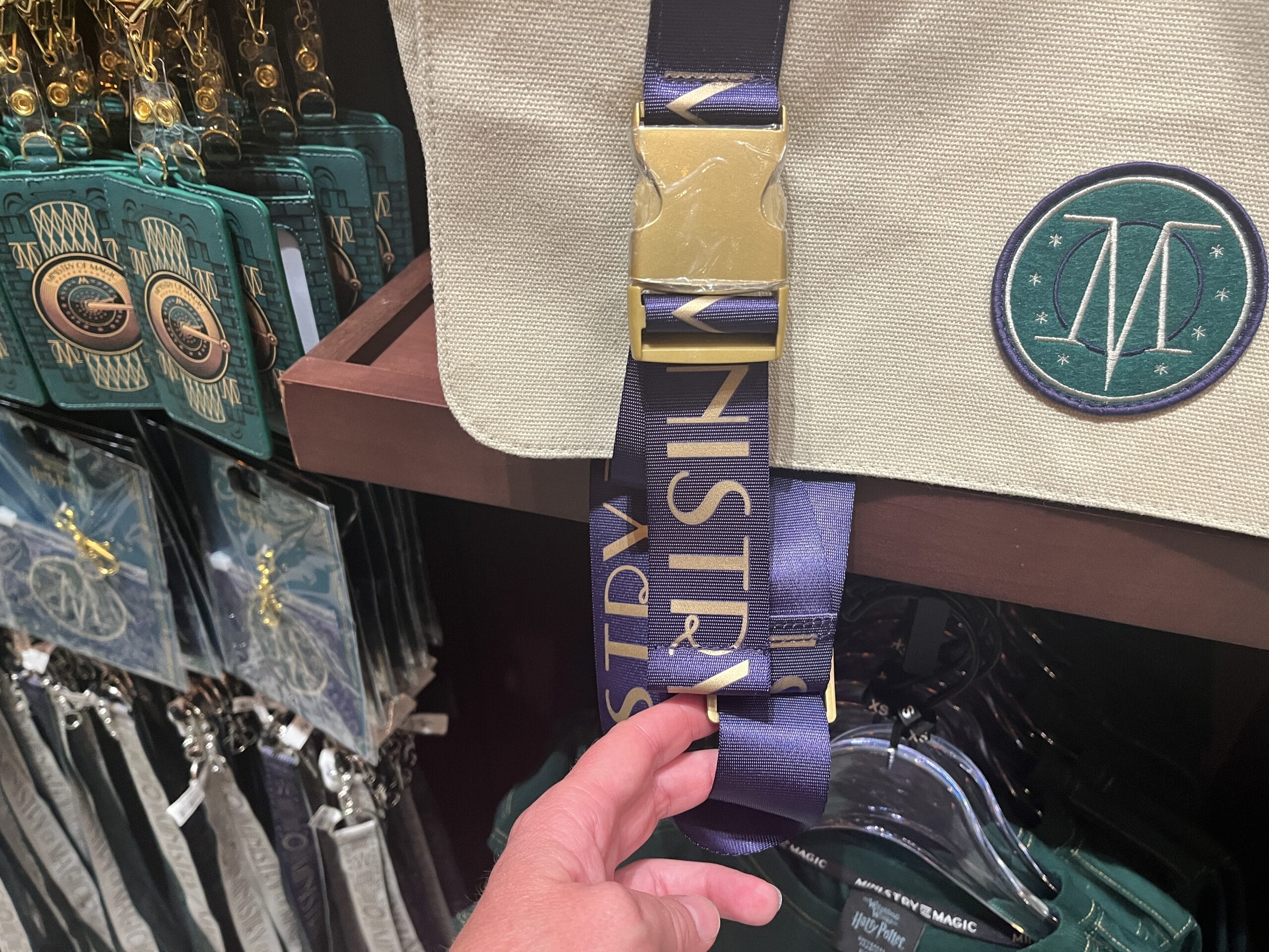 A hand holds a bag strap reading “MINISTRY” amid green and gold Wizarding World merchandise in a store.