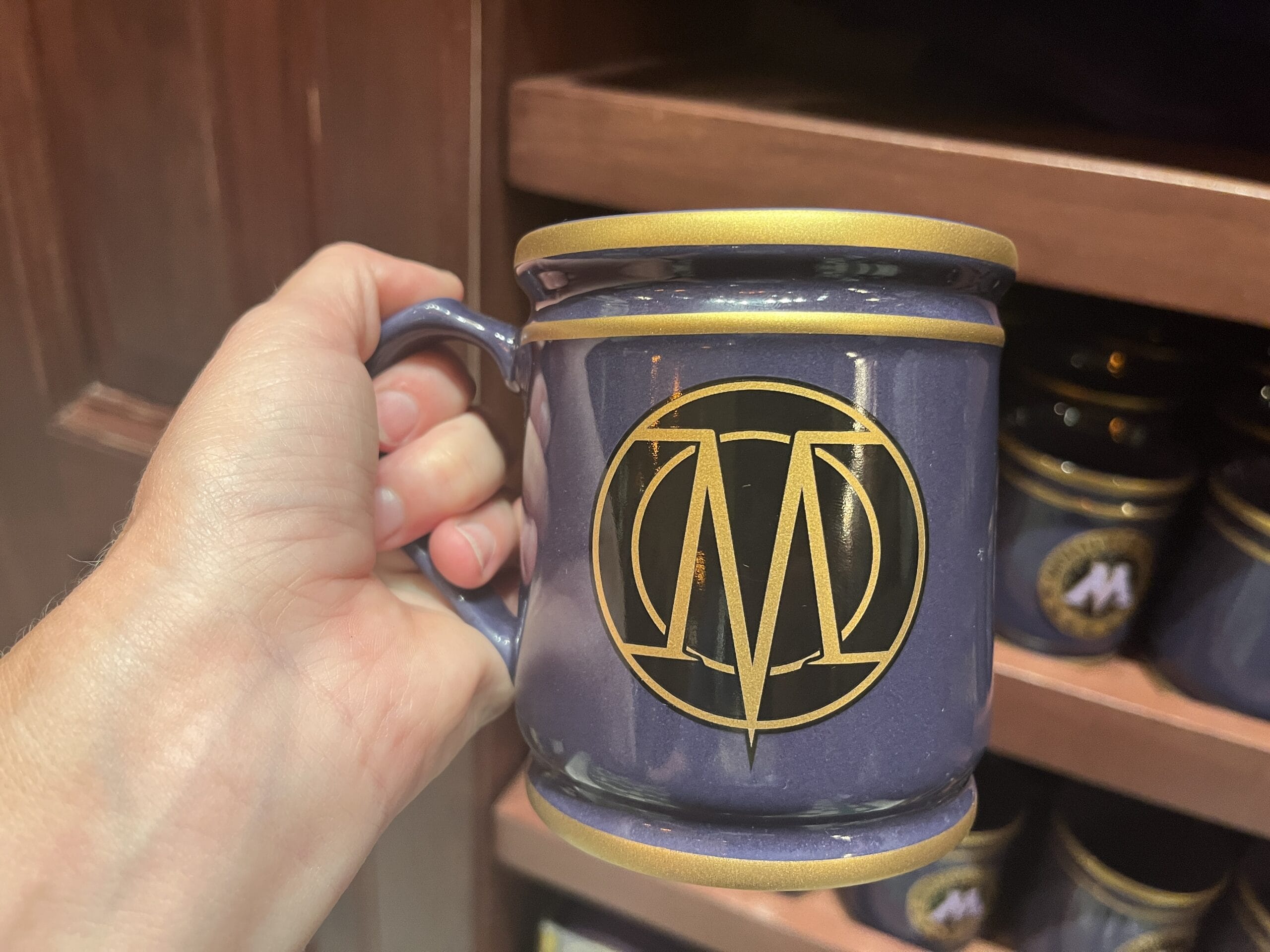 A hand holds a dark blue mug with a gold rim, sold as Epic Universe tour merchandise, featuring a gold "M" logo in a circle.