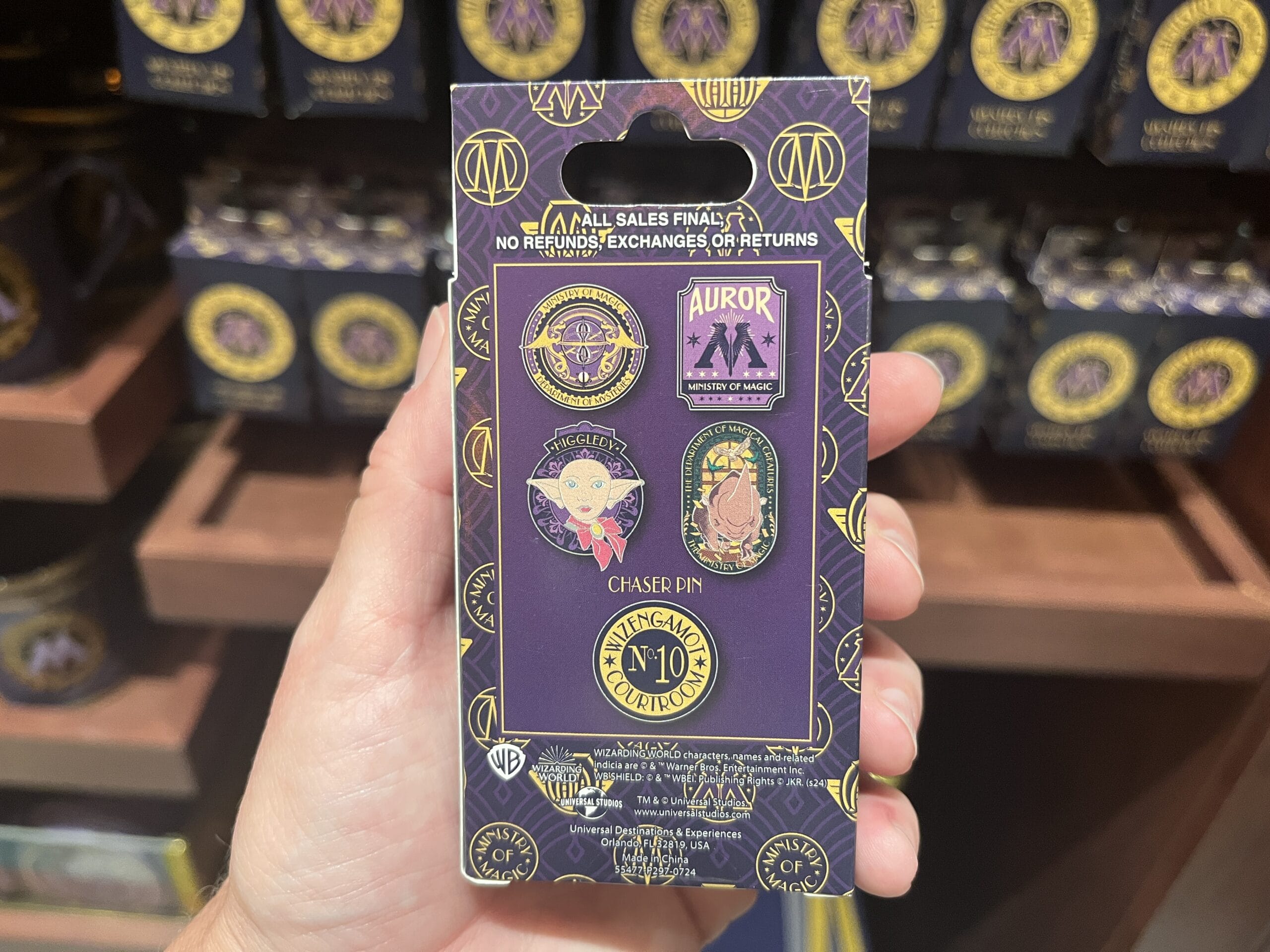 A hand holds a pack of five Wizarding World Harry Potter merchandise pins in a purple display at an Epic Universe store.