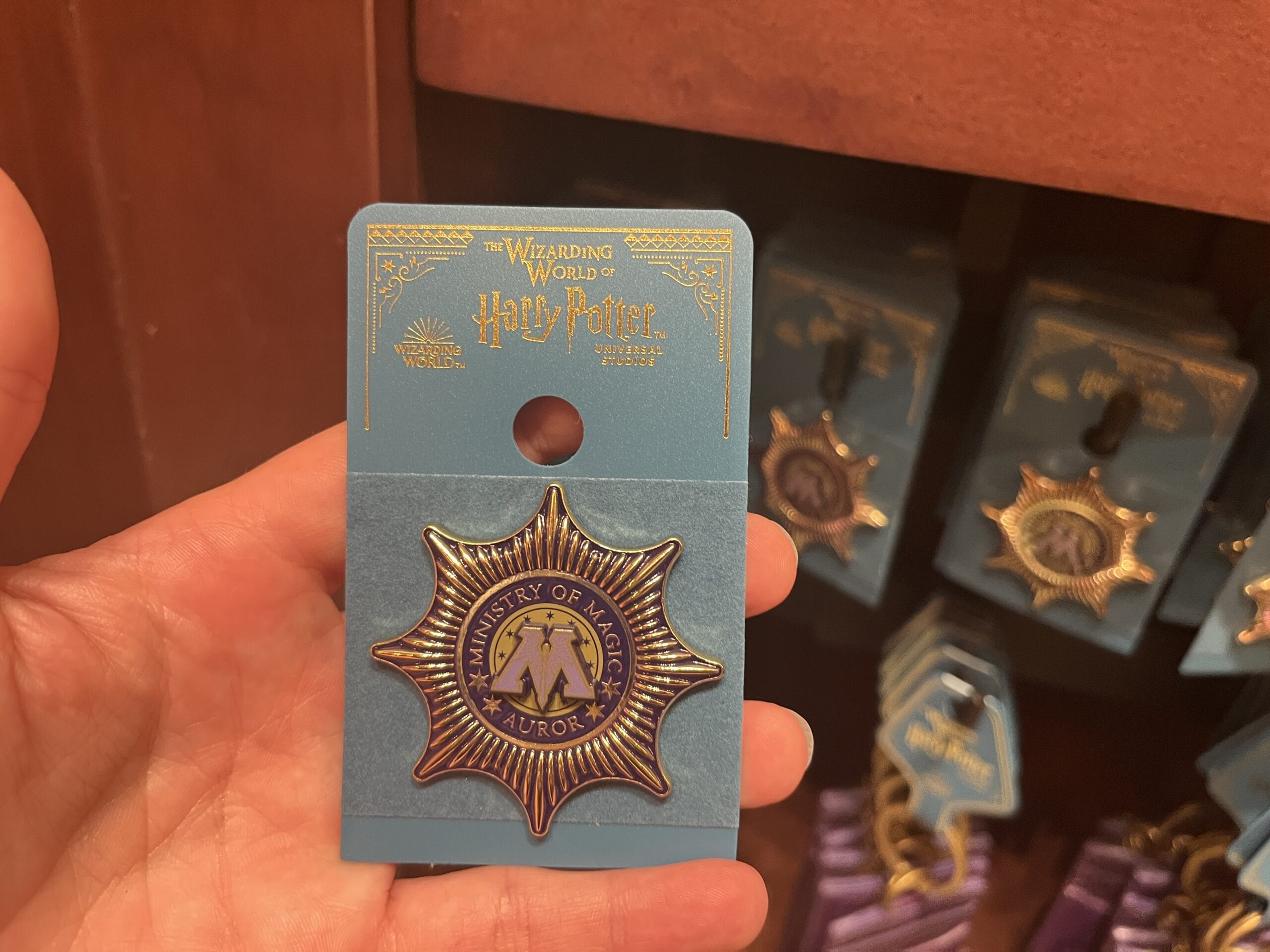 A hand holds a Ministry of Magic Auror badge, new merchandise from The Wizarding World of Harry Potter at Epic Universe.