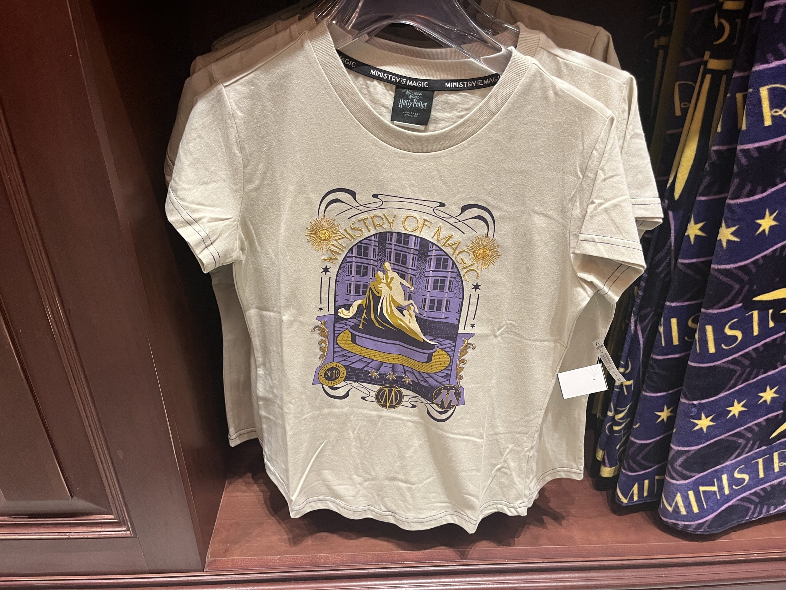 Beige T-shirt with a purple and gold "Ministry of Magic" design—perfect Wizarding World Harry Potter merchandise at Epic Universe.