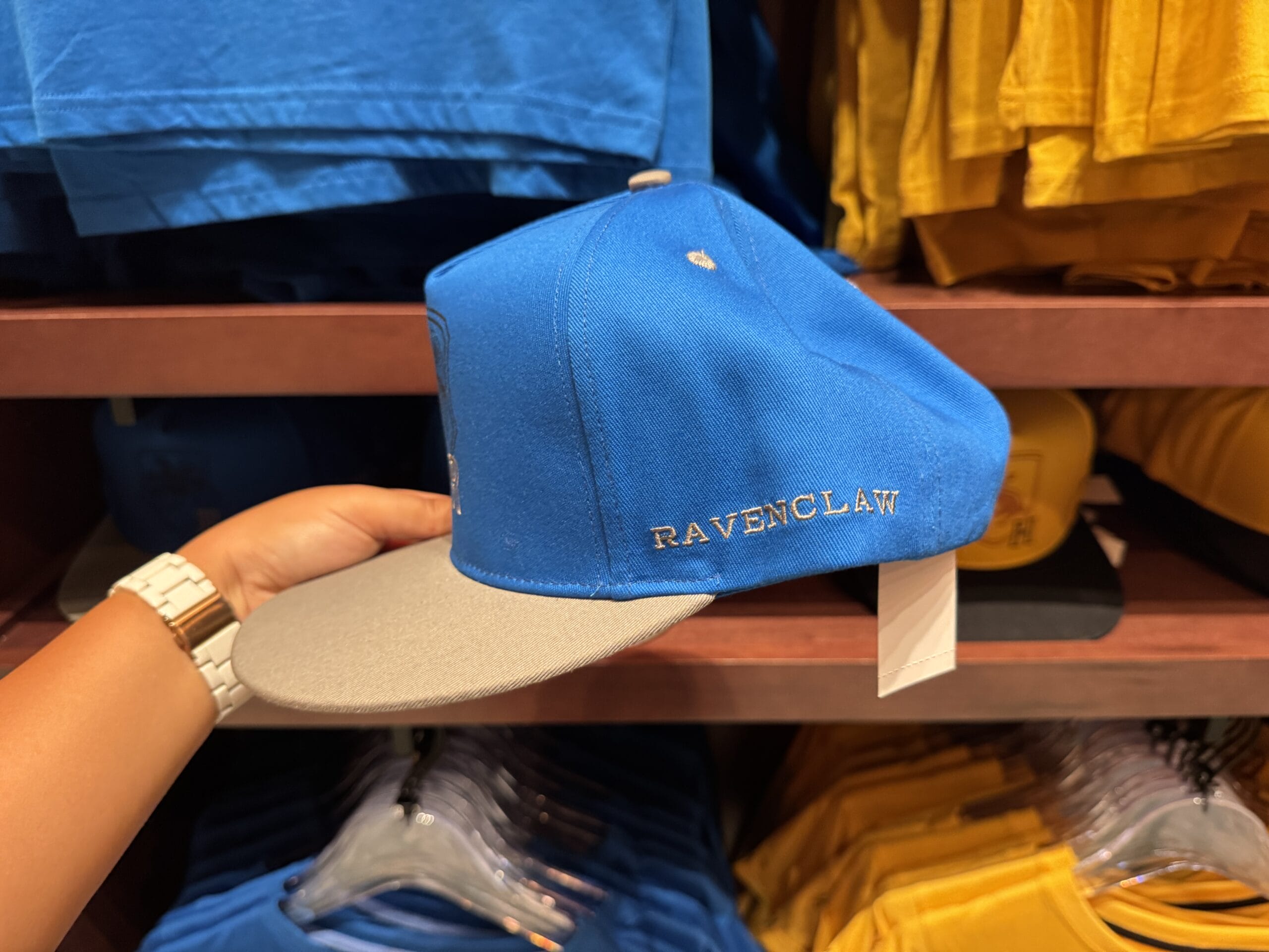 Hand holding a blue and gray "Ravenclaw" cap, surrounded by Harry Potter merchandise in a store with yellow shirts behind.