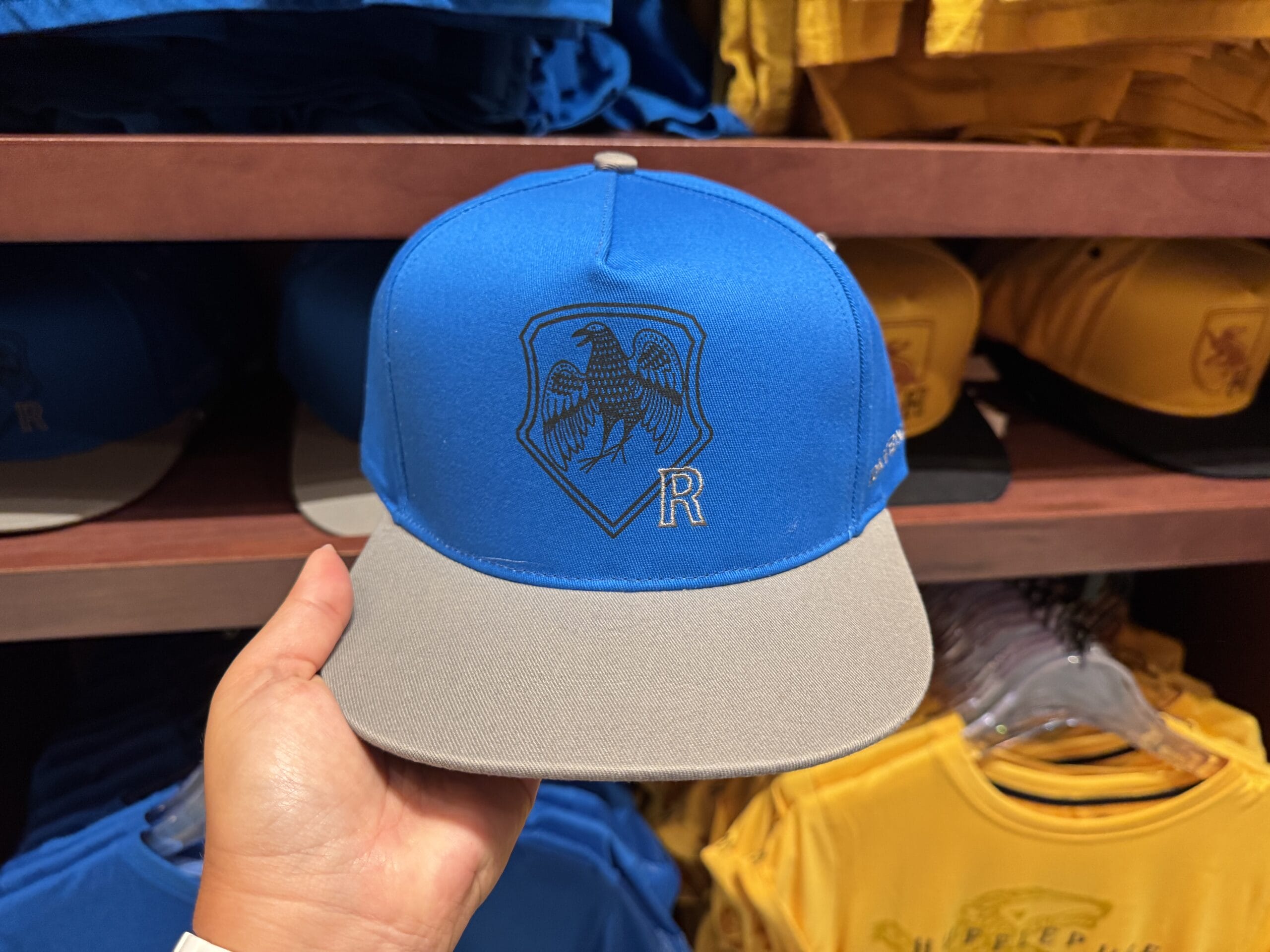 A hand holds a blue and gray cap with an eagle crest and "R" on the front, part of new Epic Universe merchandise.