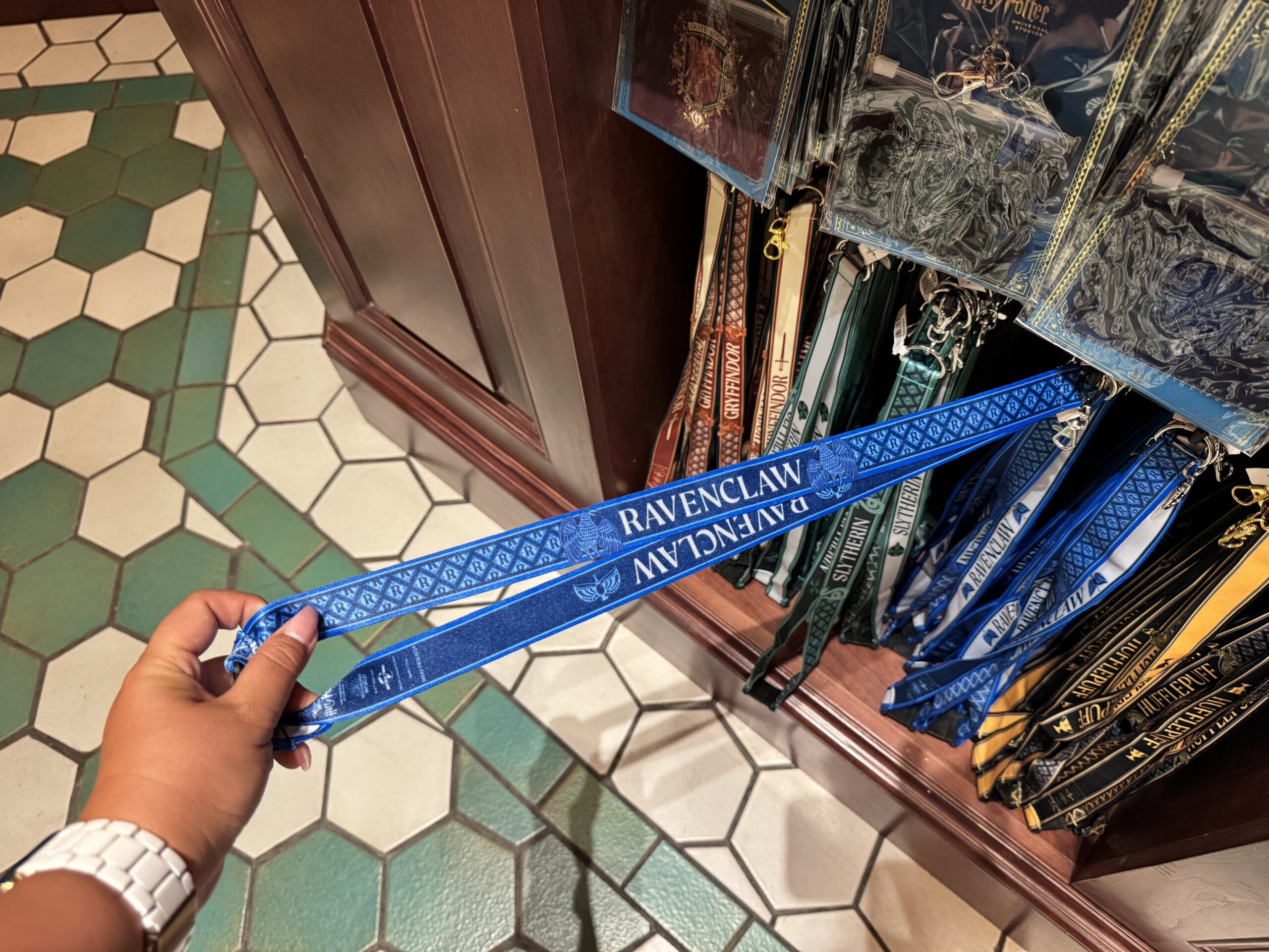 A hand holds a blue Ravenclaw lanyard among Wizarding World of Harry Potter merchandise at an Epic Universe store display.