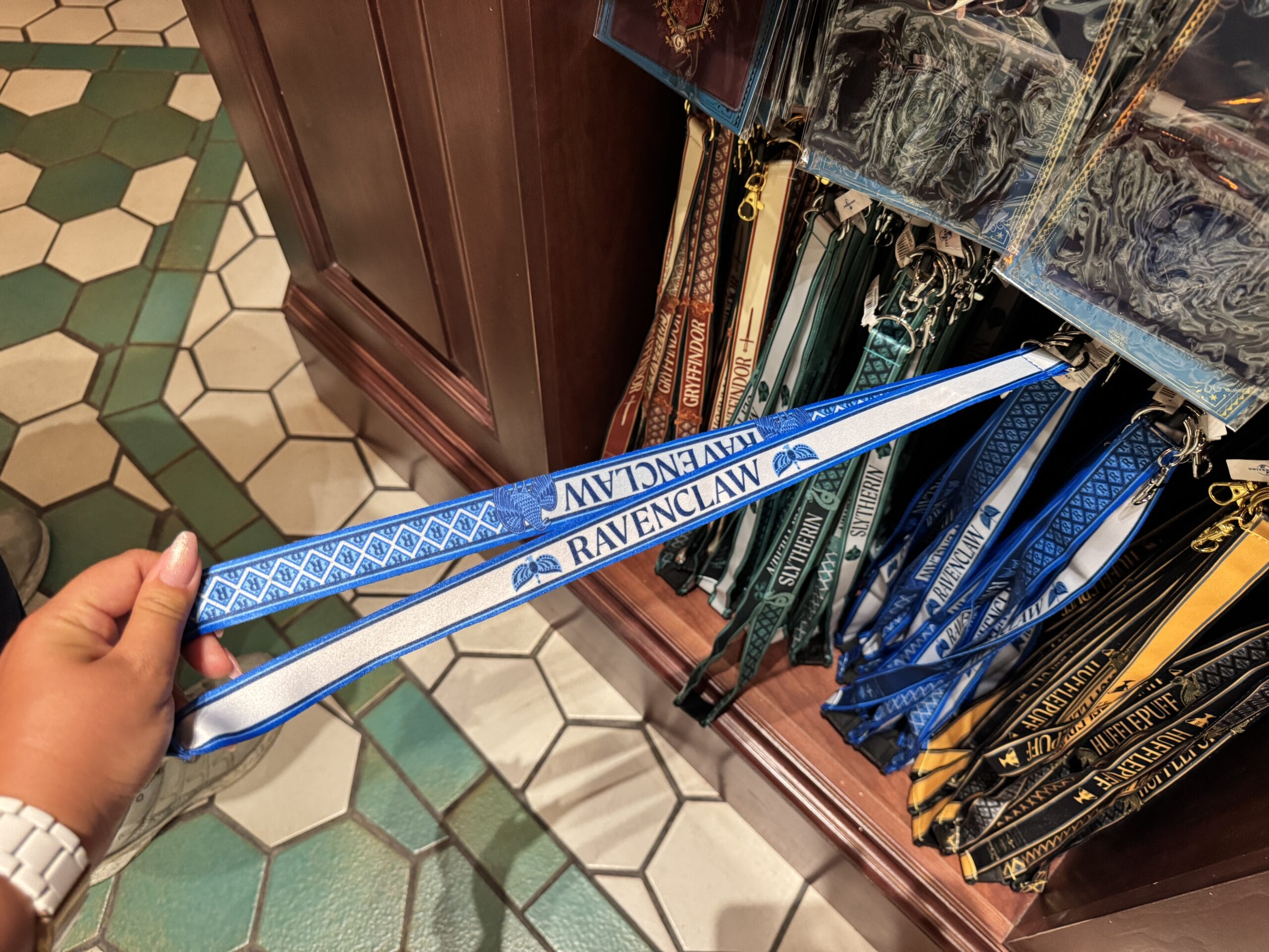 A hand holds a blue Ravenclaw lanyard near Wizarding World merchandise racks with other Hogwarts house lanyards.