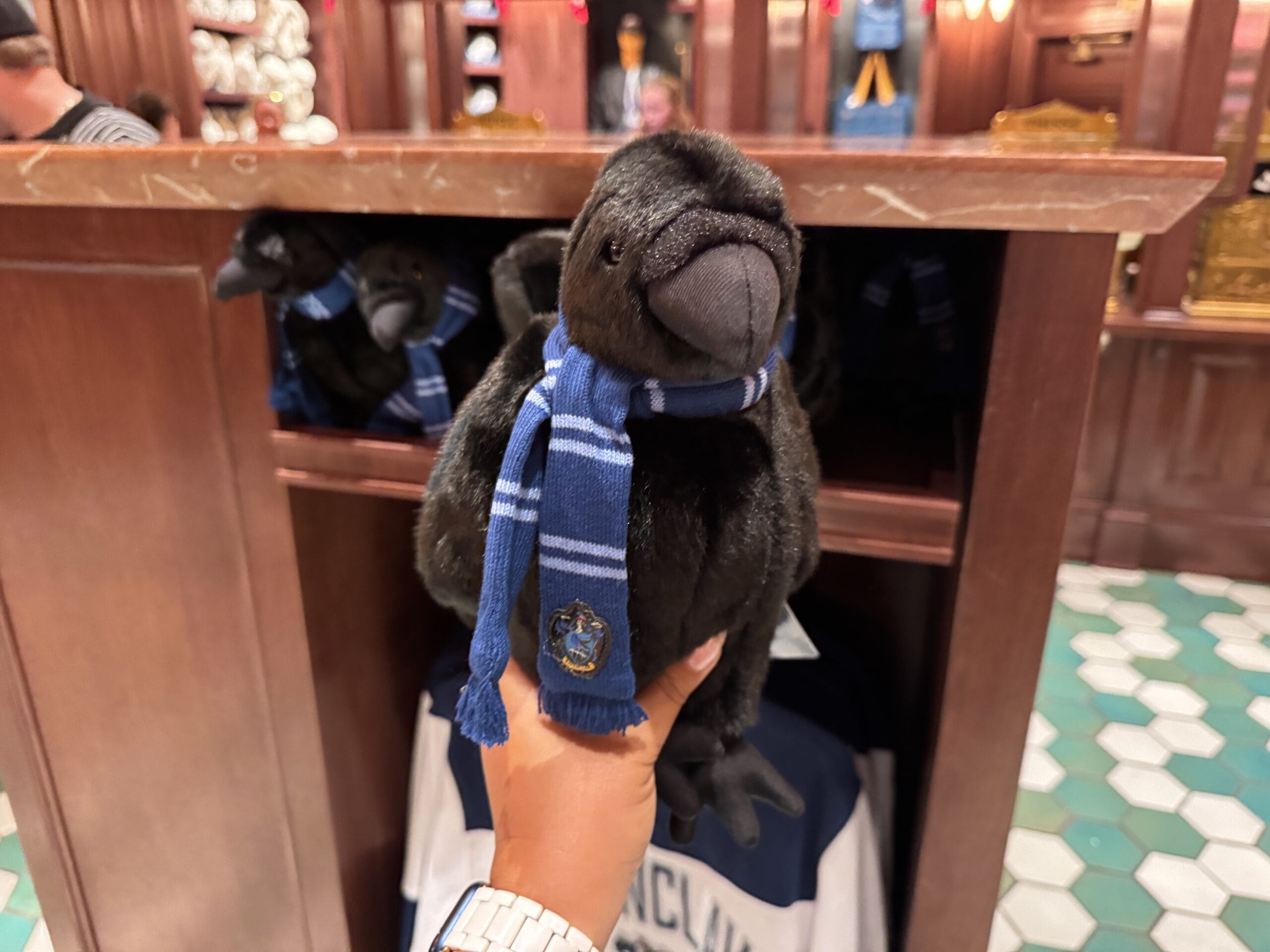 A hand holds a plush black raven in a blue Ravenclaw scarf—Epic Universe Wizarding World of Harry Potter merchandise.