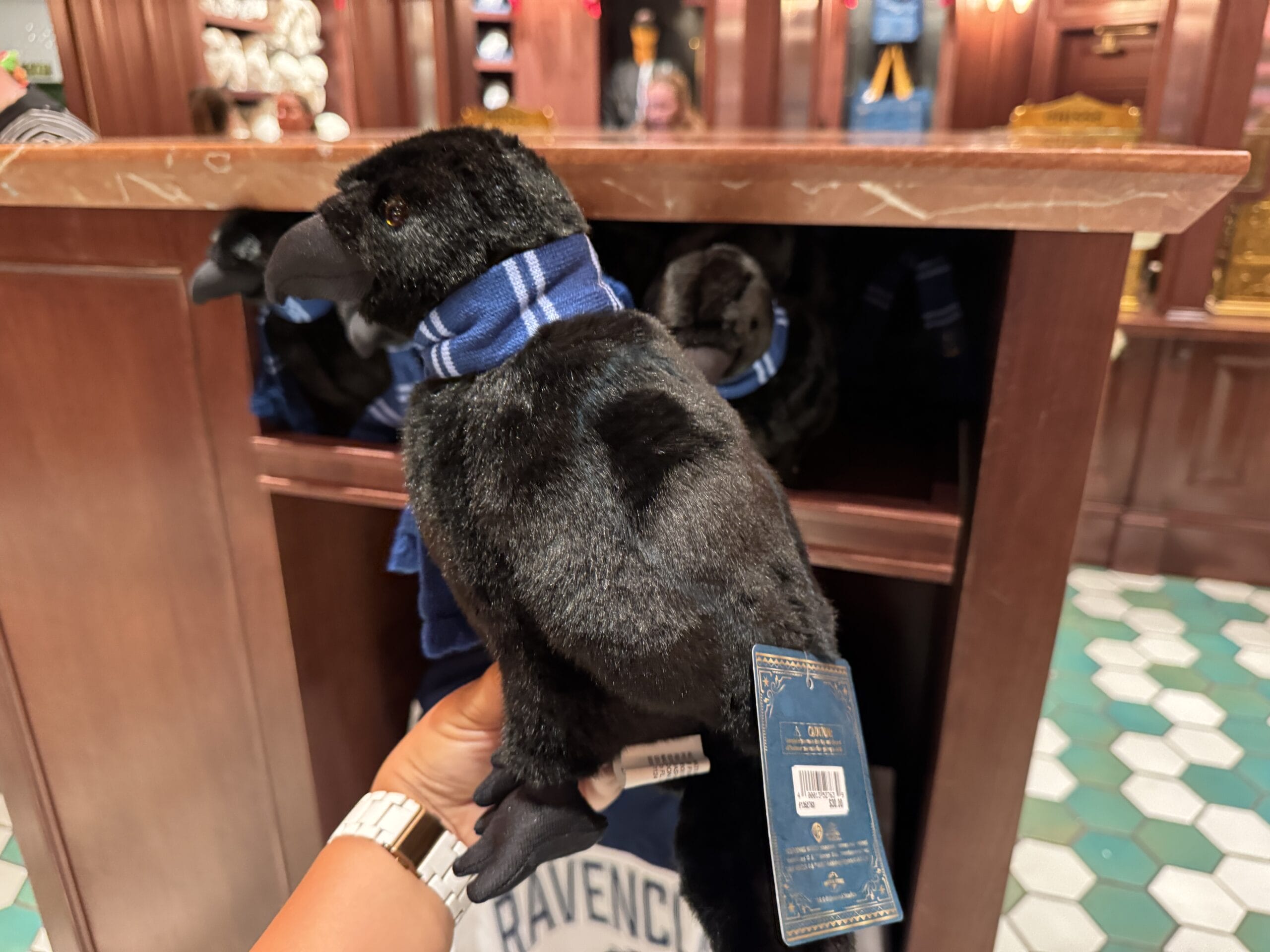 A person holds a black raven plush toy with a blue scarf, eyeing Wizarding World of Harry Potter merchandise prices in a shop.