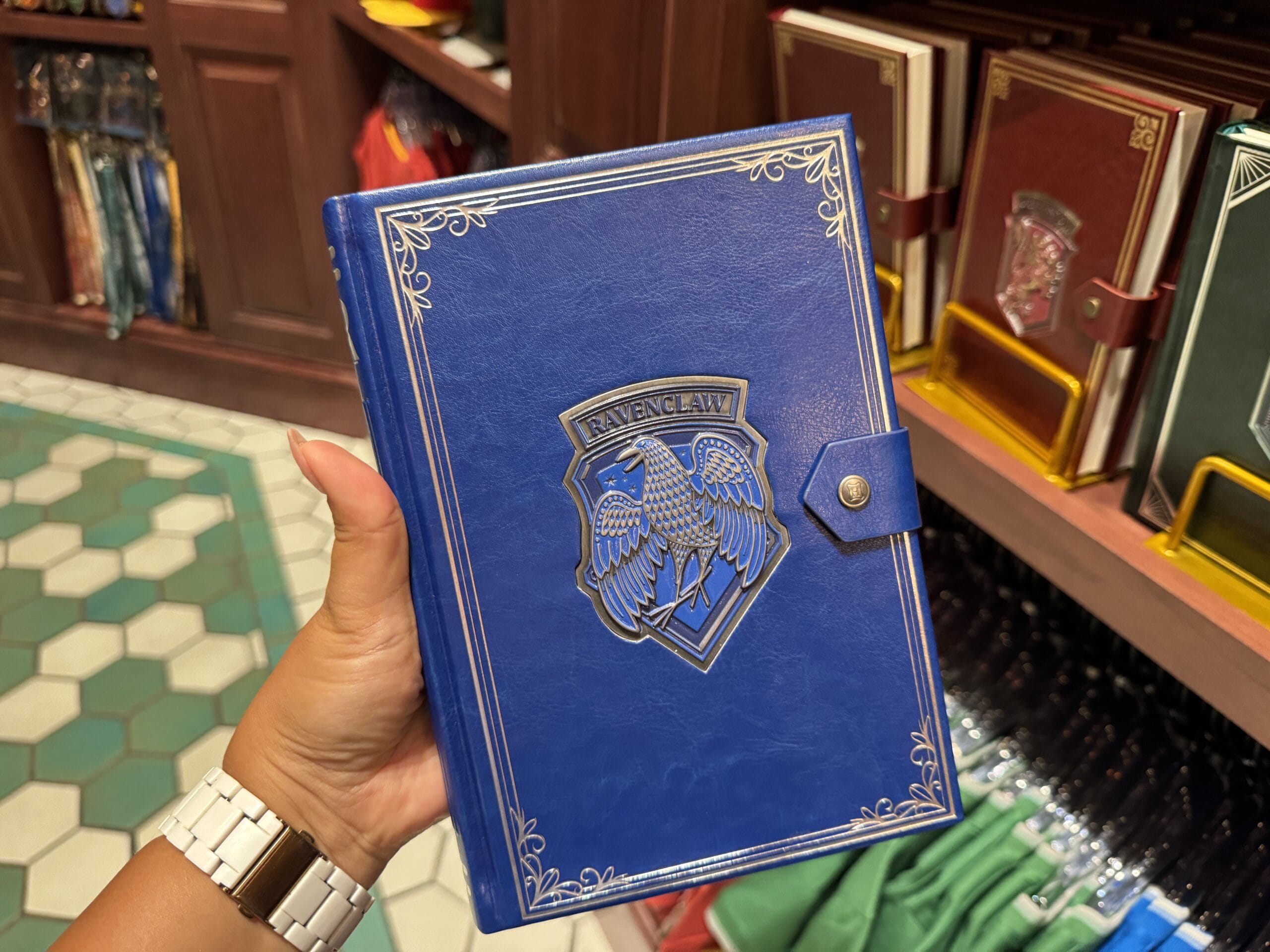 A hand holds a blue Ravenclaw notebook with a crest, displayed among Harry Potter merchandise at Epic Universe.