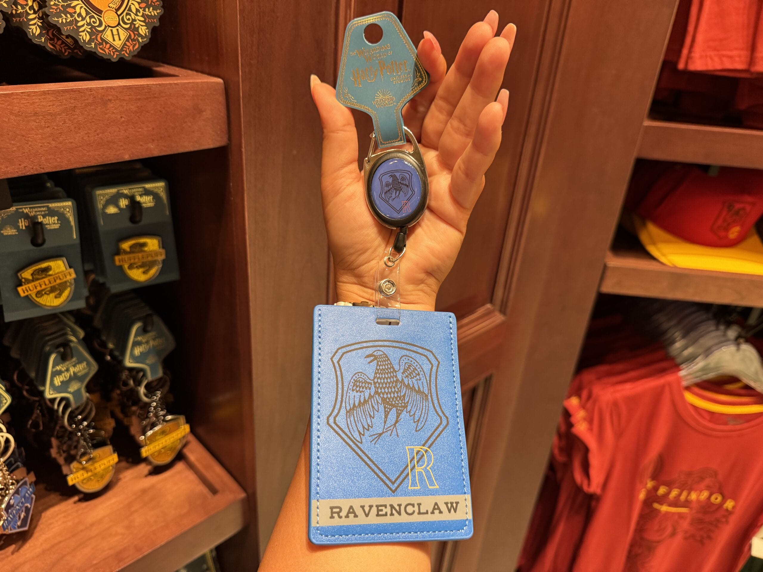A hand holds a blue Ravenclaw badge holder and keychain at the Wizarding World of Harry Potter merchandise store.
